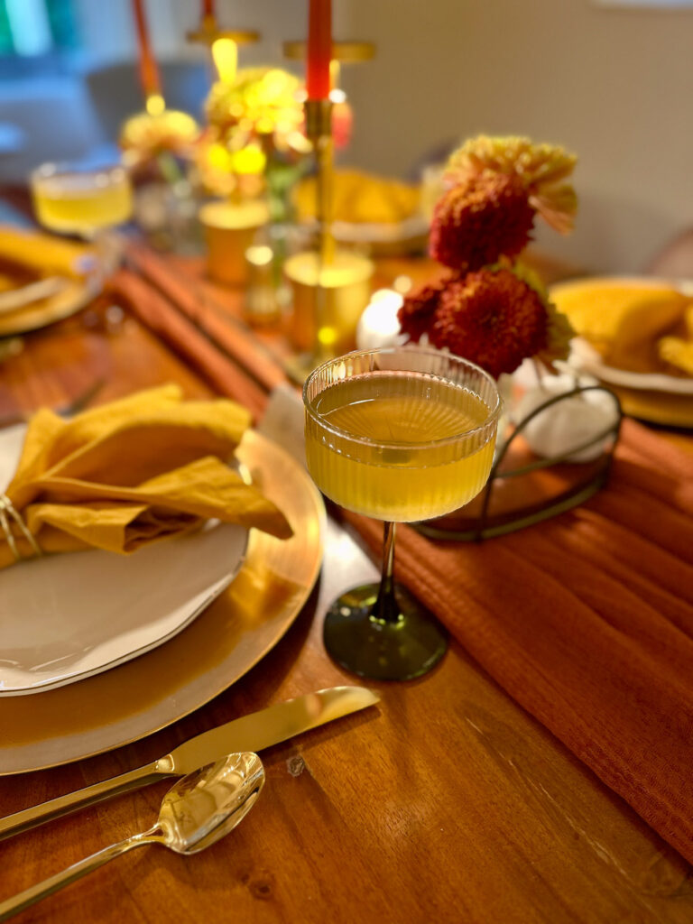 Green-stemmed coupe glass cocktail at a fall dinner party table with gold flatware, ochre napkin, and centerpiece decor.