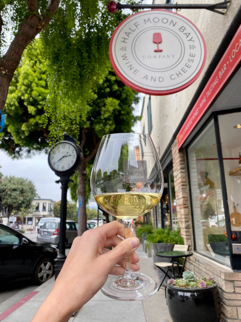 Hand holding a glass of white wine on Main Street in Half Moon Bay, with a wine shop sign and clock in the background.