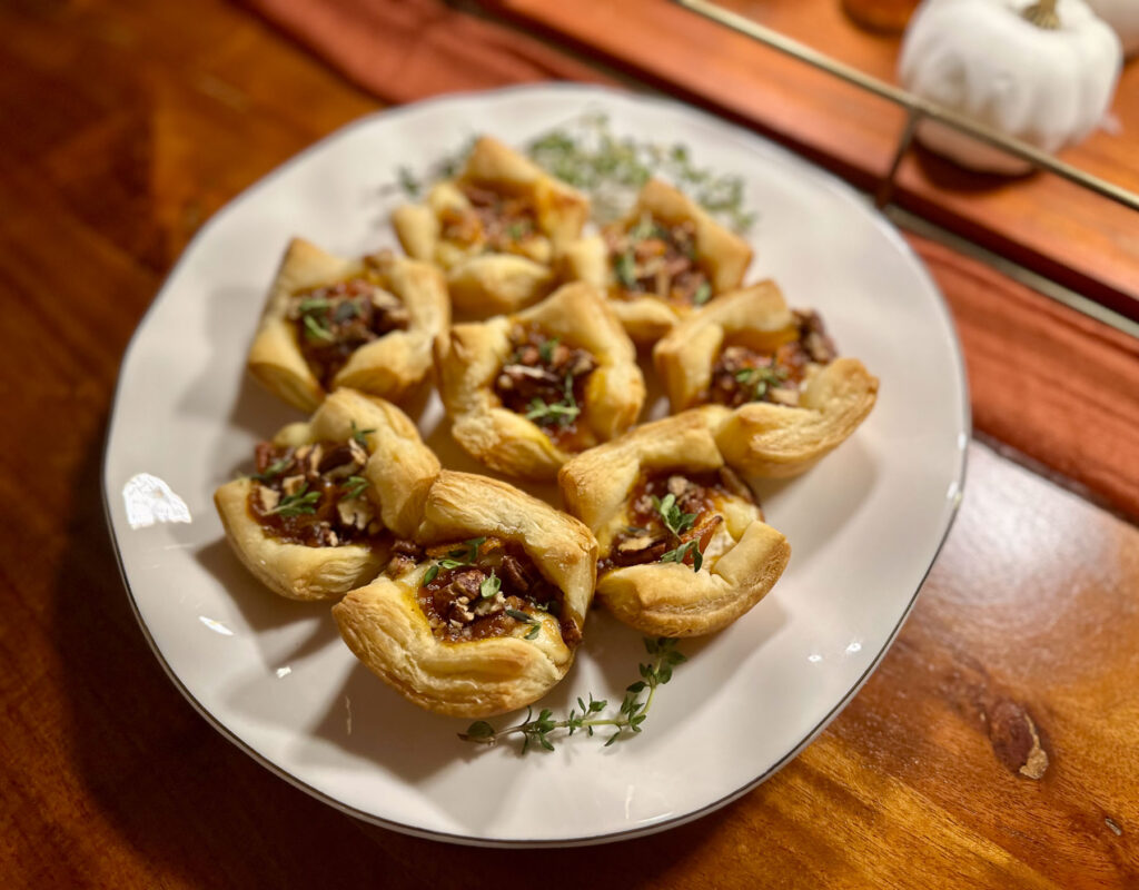 Puff pastry pumpkin butter brie bites topped with pecans and herbs on a white platter.