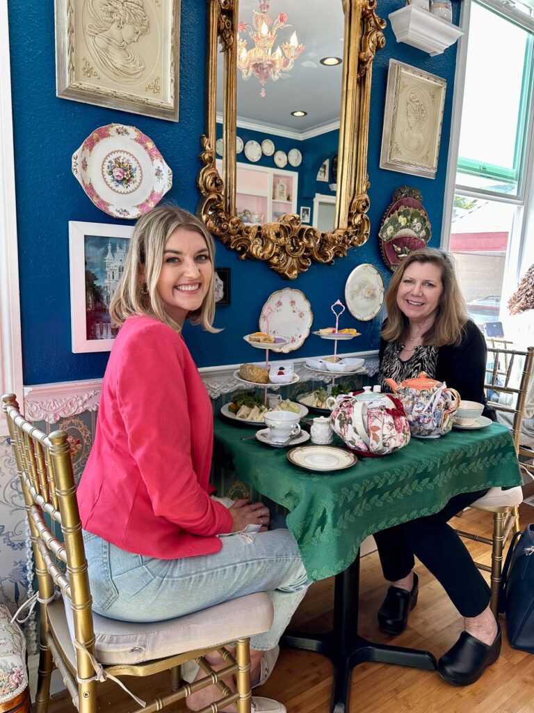 Two women enjoying afternoon tea indoors at By the Teashore, seated at a green table with tiered trays, teapots, and ornate gold-framed mirror decor.