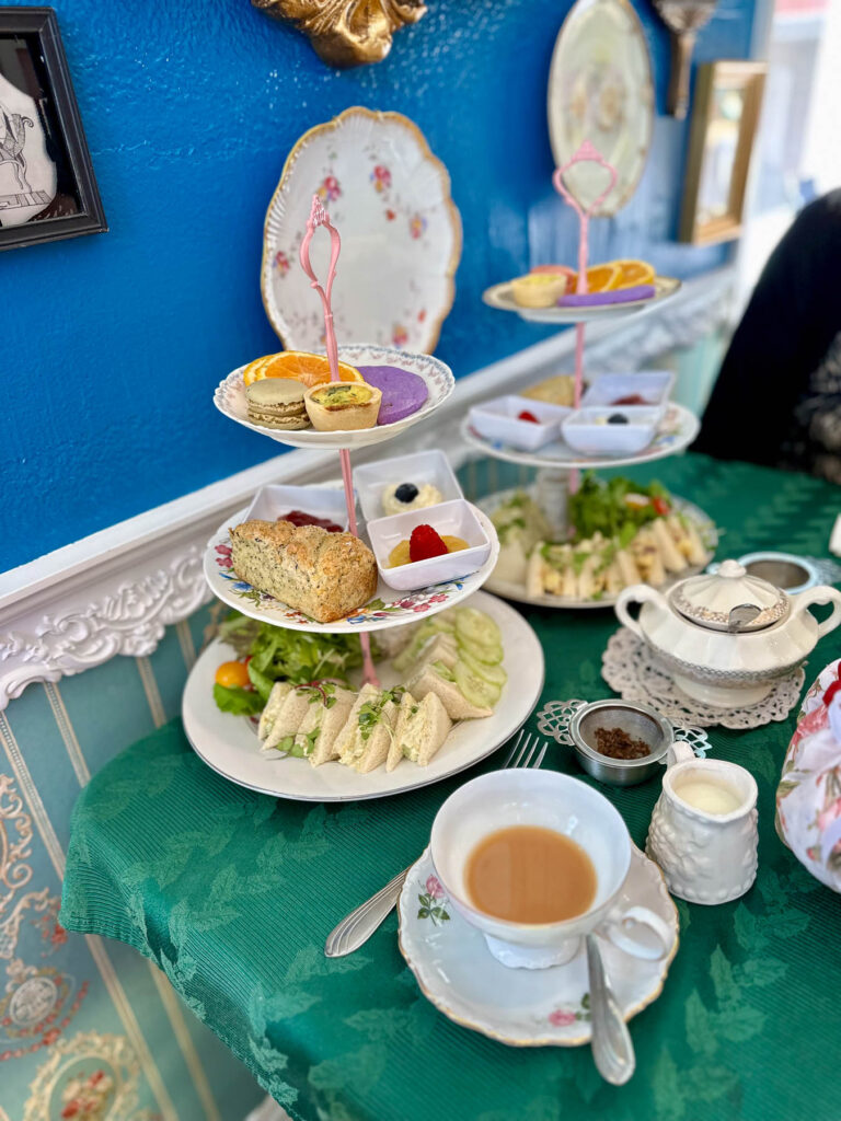 Tiered tray of afternoon tea with sandwiches, scone, quiche, and desserts, alongside a cup of tea on a green tablecloth.