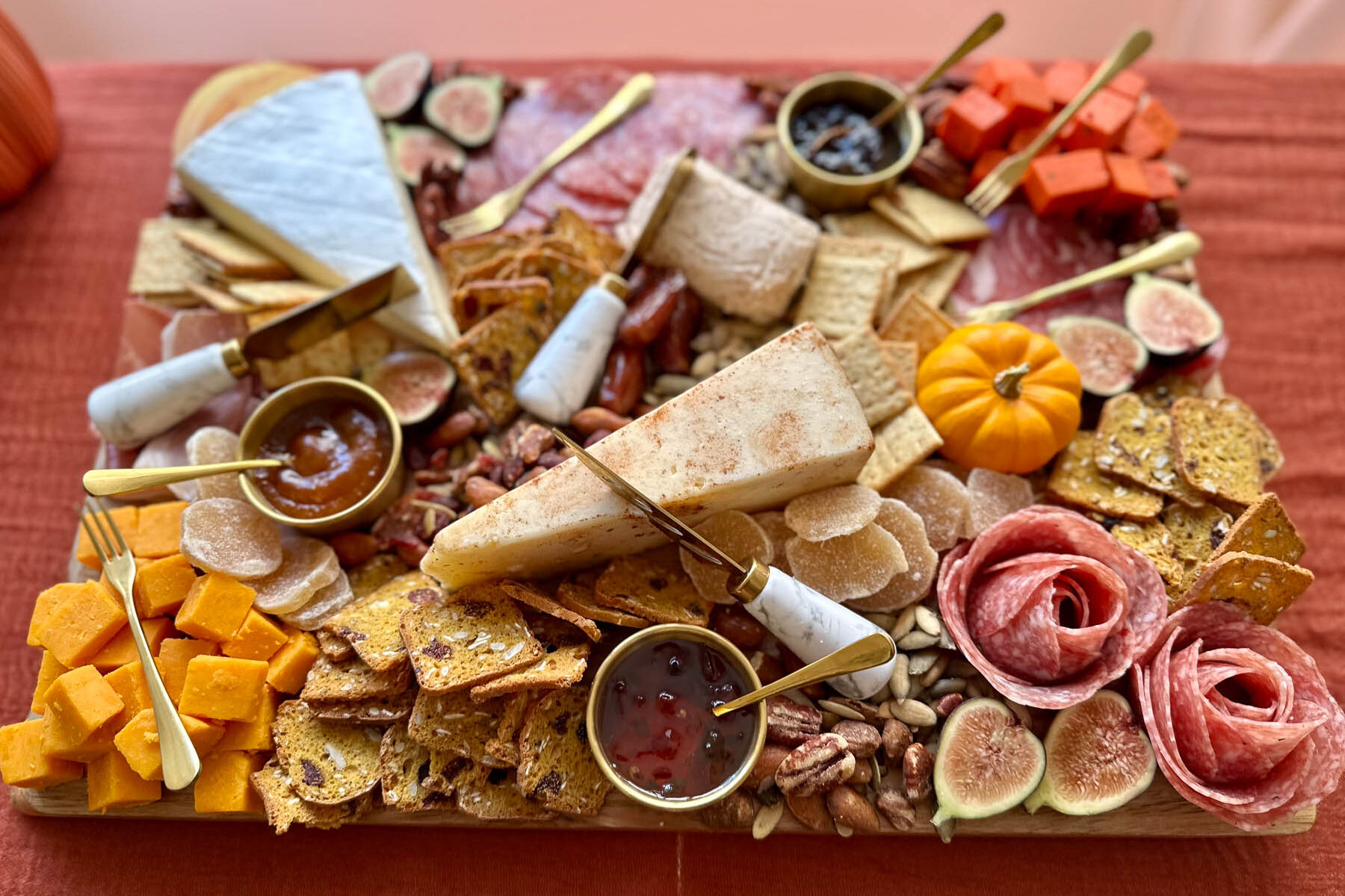 Trader Joe’s fall charcuterie board filled with cheeses, crackers, dips in gold ramekins, fresh figs, Honeycrisp apple slices, candied ginger, dates, nuts, salami roses, and mini pumpkins, arranged on a wood board with gold utensils.