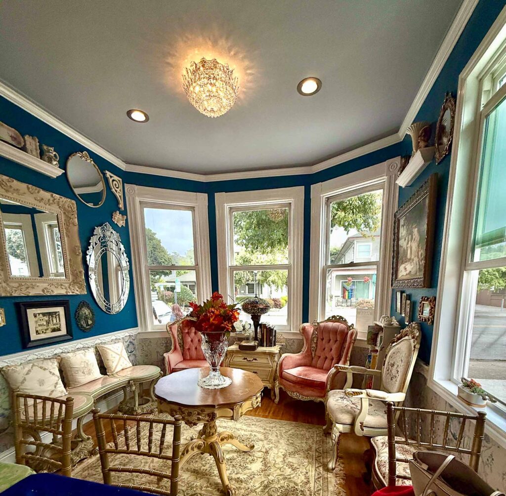 Bright corner seating area at By the Teashore with teal walls, vintage mirrors, chandeliers, and pink tufted chairs arranged around a round table.