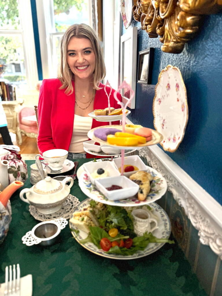 Woman smiling at a table set for afternoon tea, with tiered trays and a bright teal wall behind.