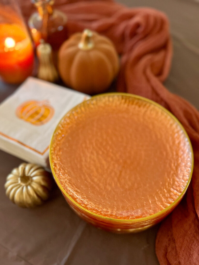 A close-up details shot of a cozy fall party setup showing a stack of textured orange plates, a pumpkin-printed paper napkin, and a tiny gold decorative pumpkin.