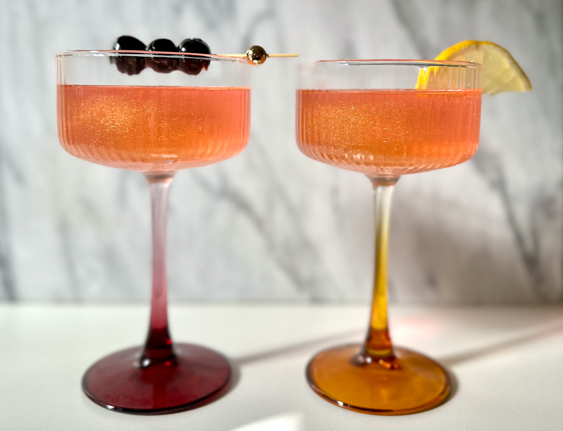Two sparkling pink drinks in coupe glasses — one garnished with cherries, the other with a lemon slice — representing cocktails and mocktails in a side-by-side comparison.