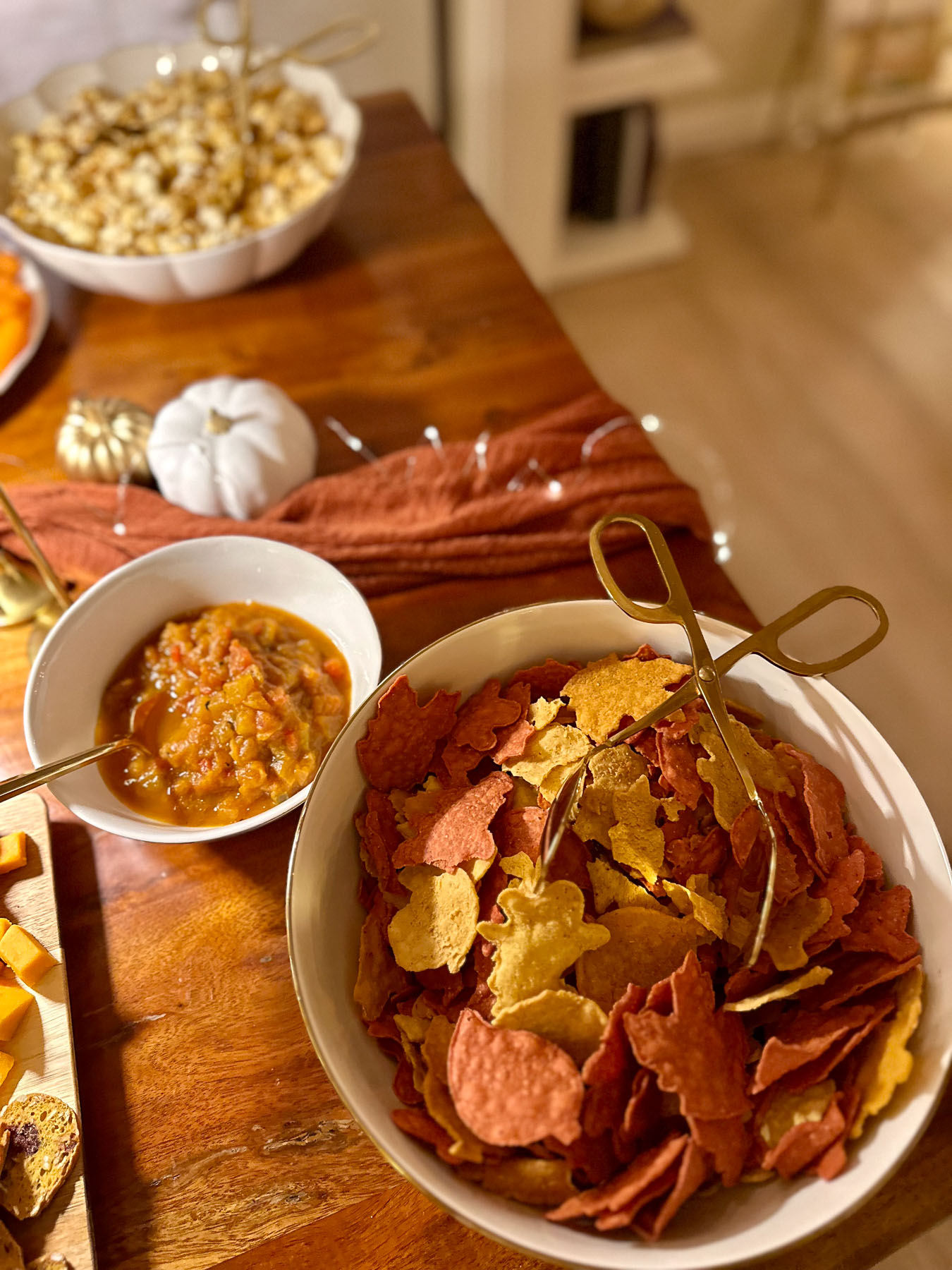 A white bowl filled with festive orange, yellow, and red leaf-shaped tortilla chips next to a small bowl of harvest salsa and a plate of fresh carrots.