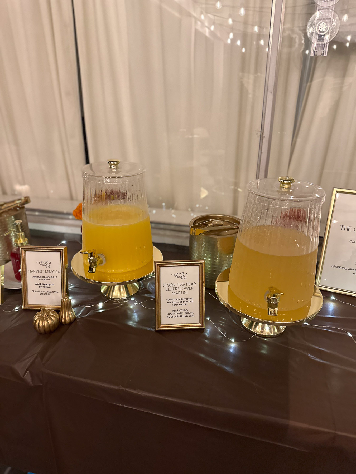 Two clear, ribbed drink dispensers filled with cocktails on a dark tabletop bar setup with gold-framed cocktail menu signs.