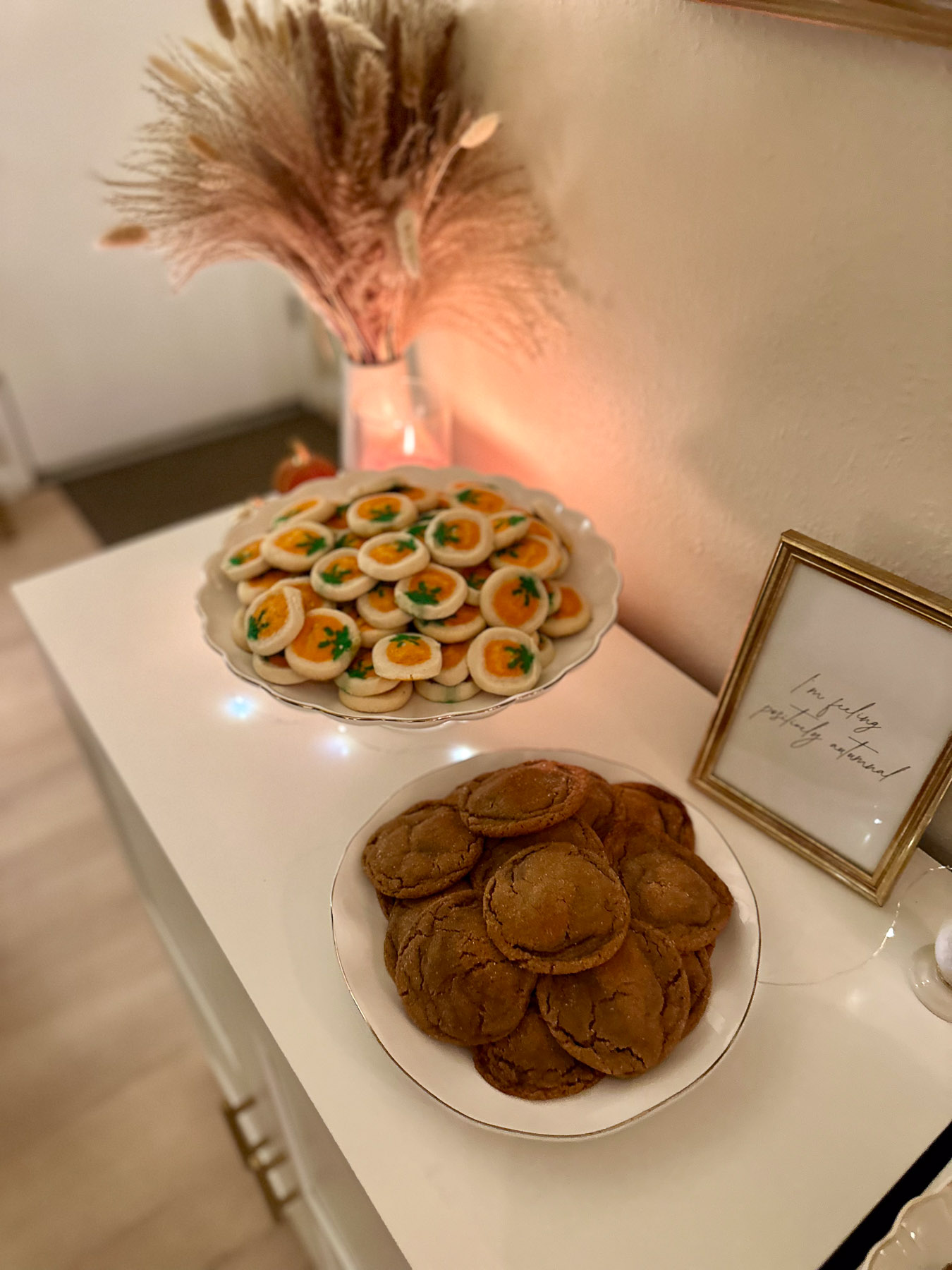 Platters of homemade party desserts on a white cabinet, including a stack of old-fashioned molasses cookies and a raised plate of cutesy pumpkin-spice sugar cookies.