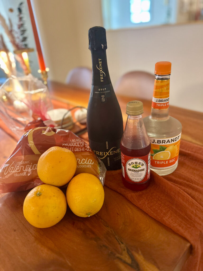 Ingredients for Halloween Harvest Mimosas — fresh Valencia oranges, Freixenet cava, Rose’s grenadine, and triple sec displayed on a fall table runner.