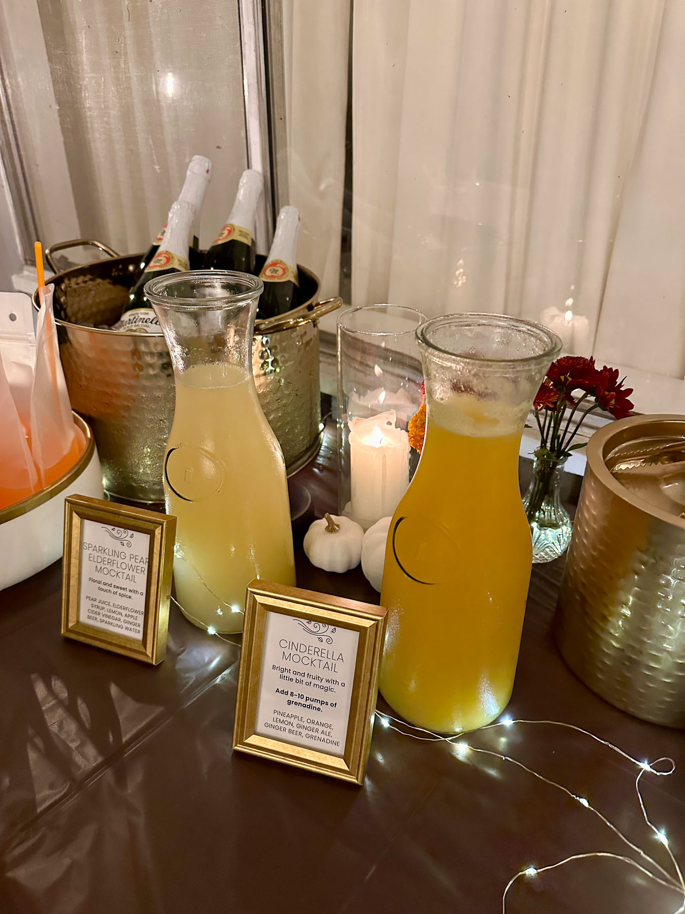 A self-serve party bar featuring glass carafes filled with yellow mocktails, a gold bucket of Martinelli's bottles, and small gold-framed menus.