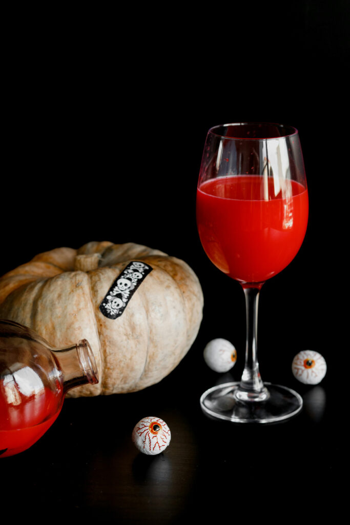 Red Halloween cocktail in a wine glass with a white pumpkin and fake eyeballs on a dark backdrop — a spooky yet stylish drink for hosting a Halloween party.