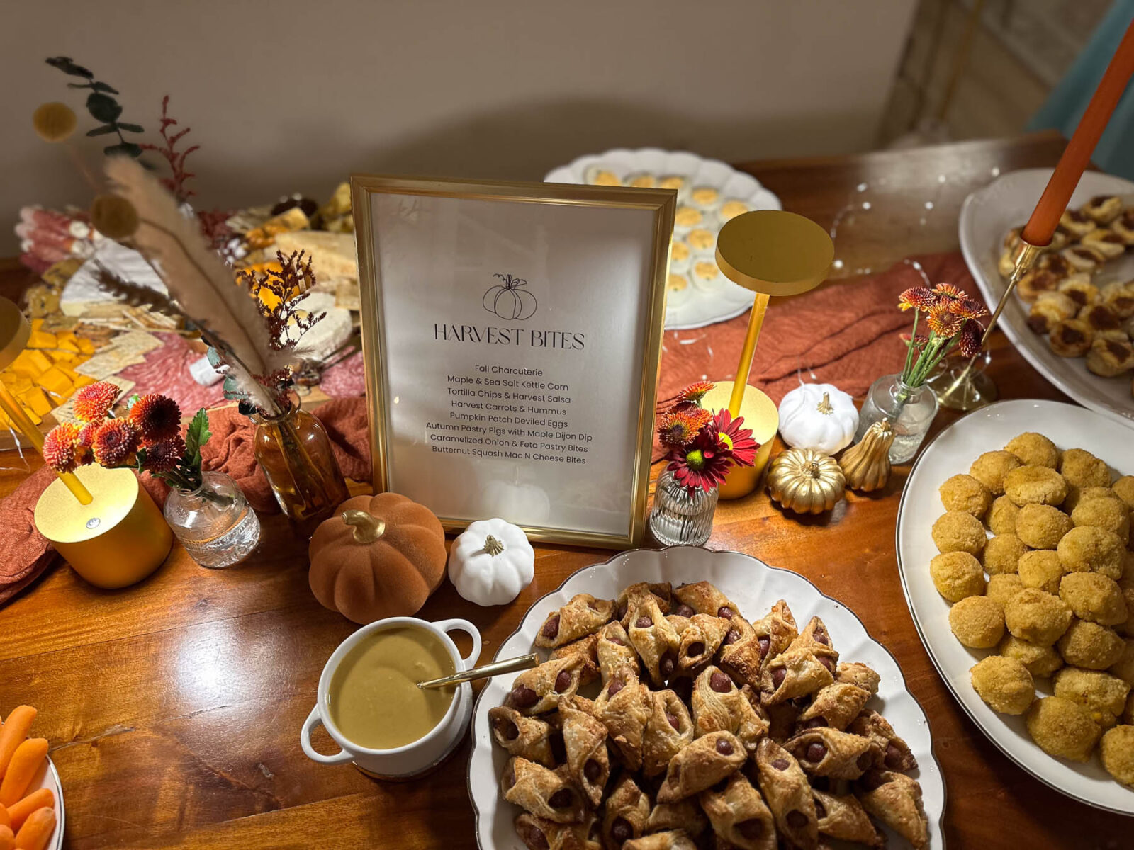 A wooden table set with Halloween party food, featuring a framed 'Harvest Bites' menu, mini pumpkins, pastry pigs in a blanket, and a plate of fried mac and cheese bites.