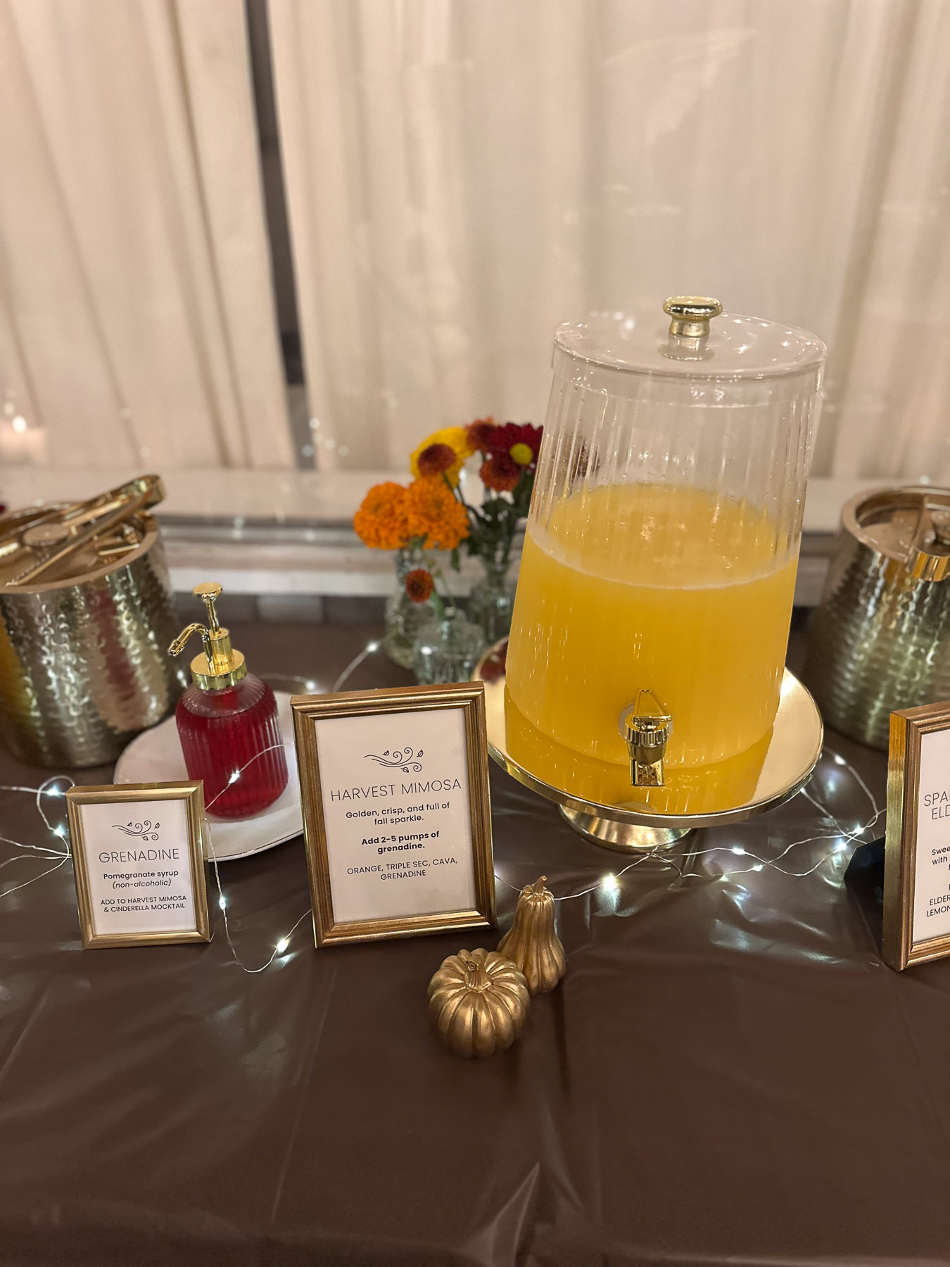 A clear drink dispenser filled with a festive orange harvest mimosa cocktail next to a small bottle of grenadine syrup, gold pumpkins, and fairy lights.