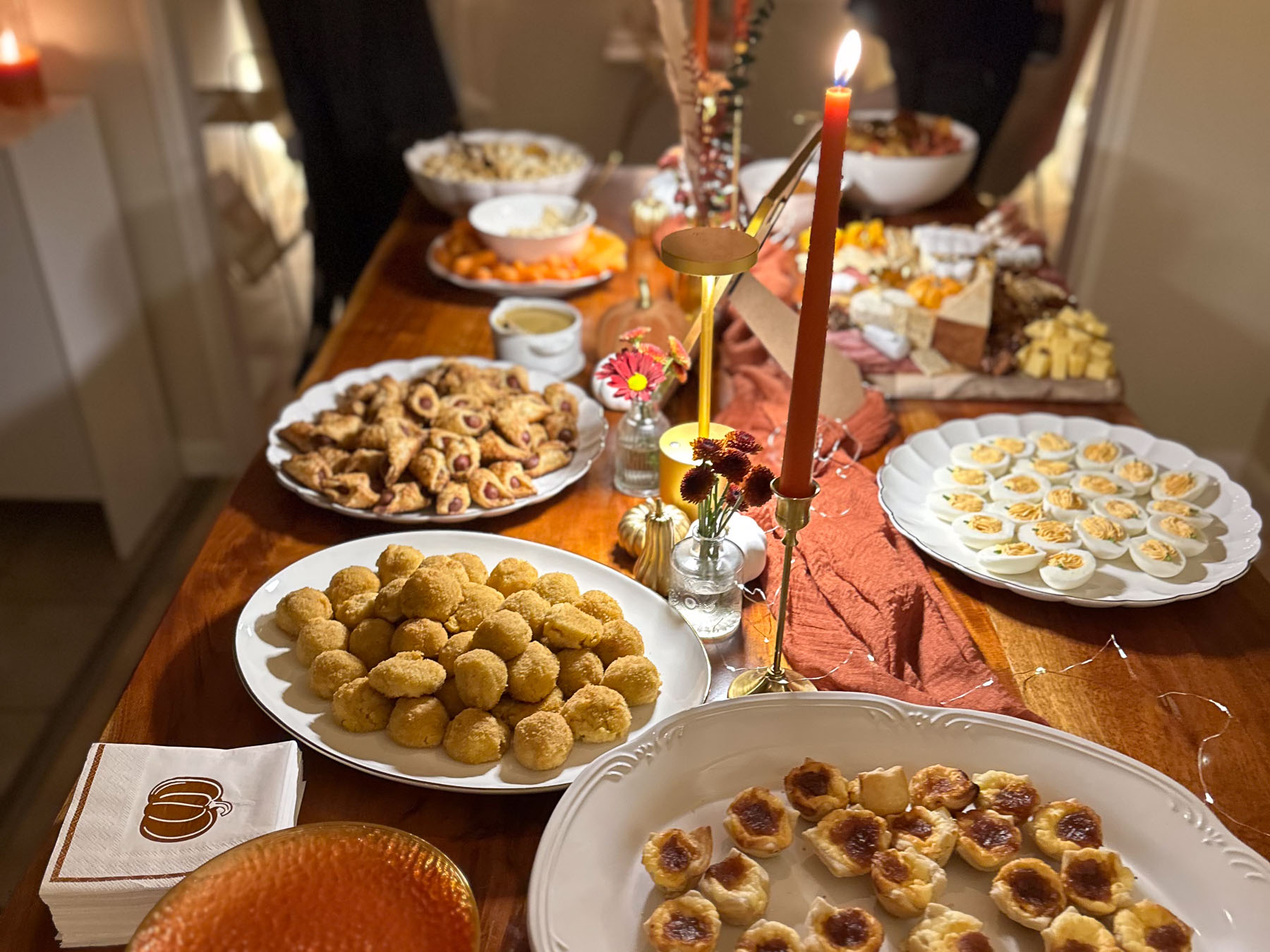 A cozy dining table set for hosting a halloween party, featuring a lit orange taper candle in the foreground, mini decorative pumpkins, and large white platters filled with pastry bites, deviled eggs, and charcuterie on a rust-colored runner.