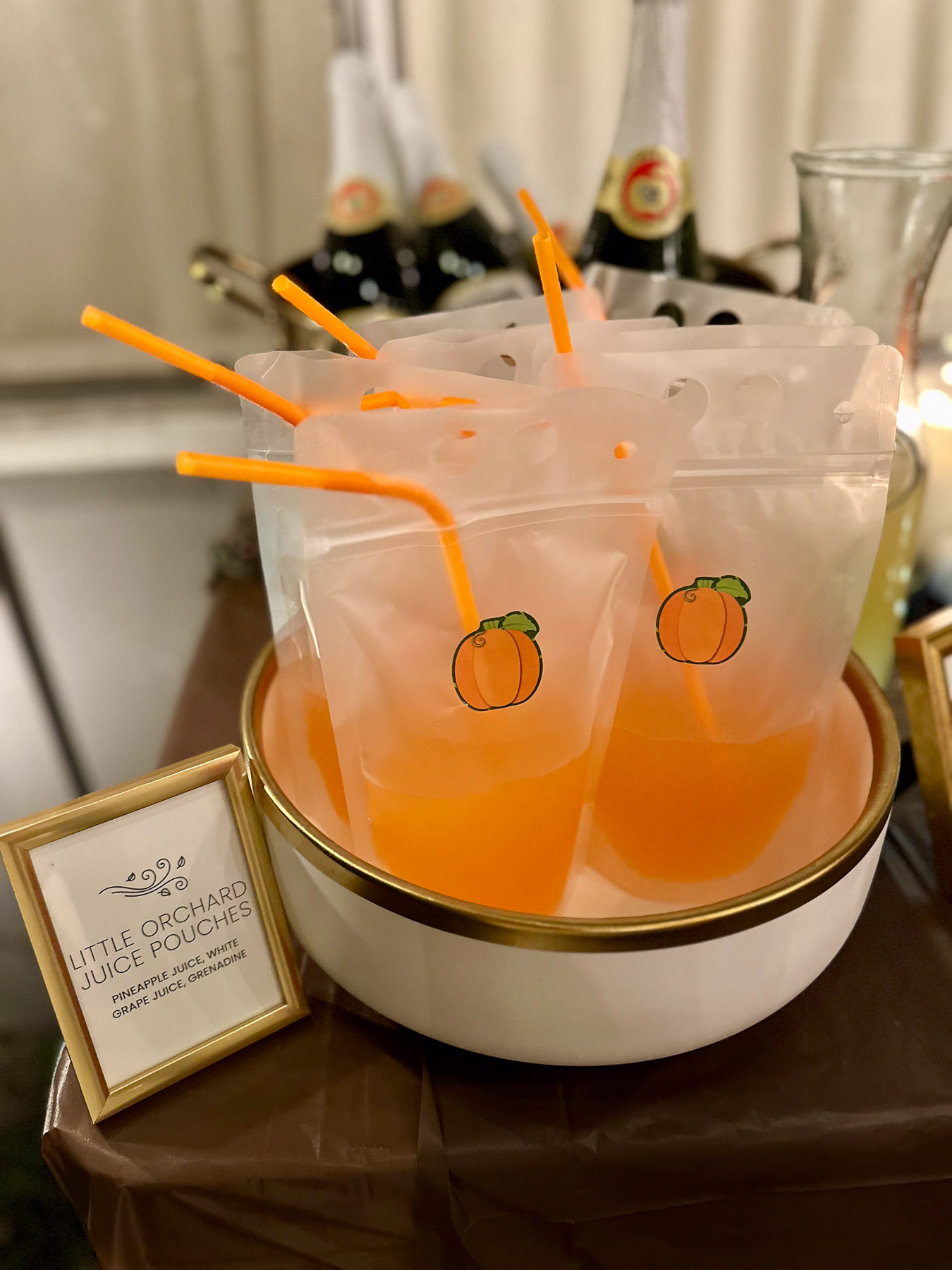A white bowl filled with clear, reusable drink pouches with orange straws and pumpkin stickers, next to a gold-framed sign that reads 'Little Orchard Juice Pouches'.