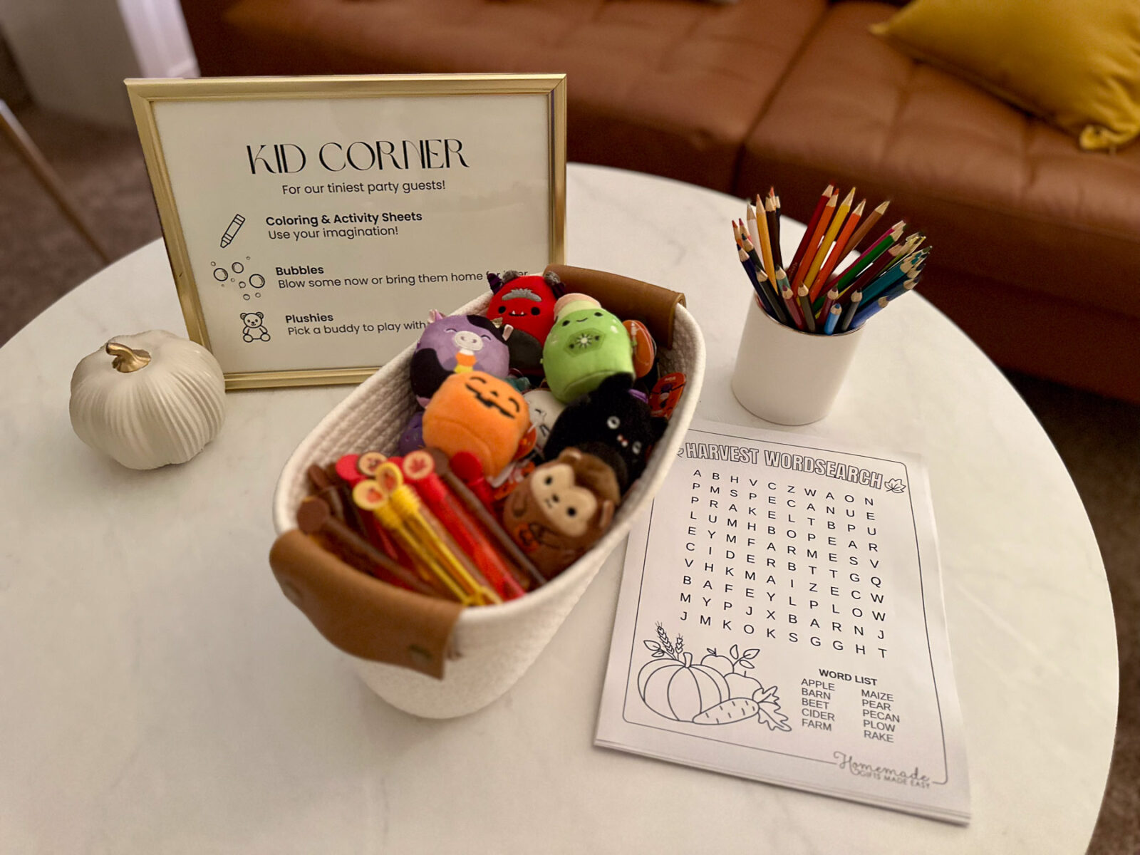 A designated 'Kid Corner' on a white round table featuring a basket of mini Halloween plushies, mini bubble wands, a jar of colored pencils, and a harvest word search puzzle.
