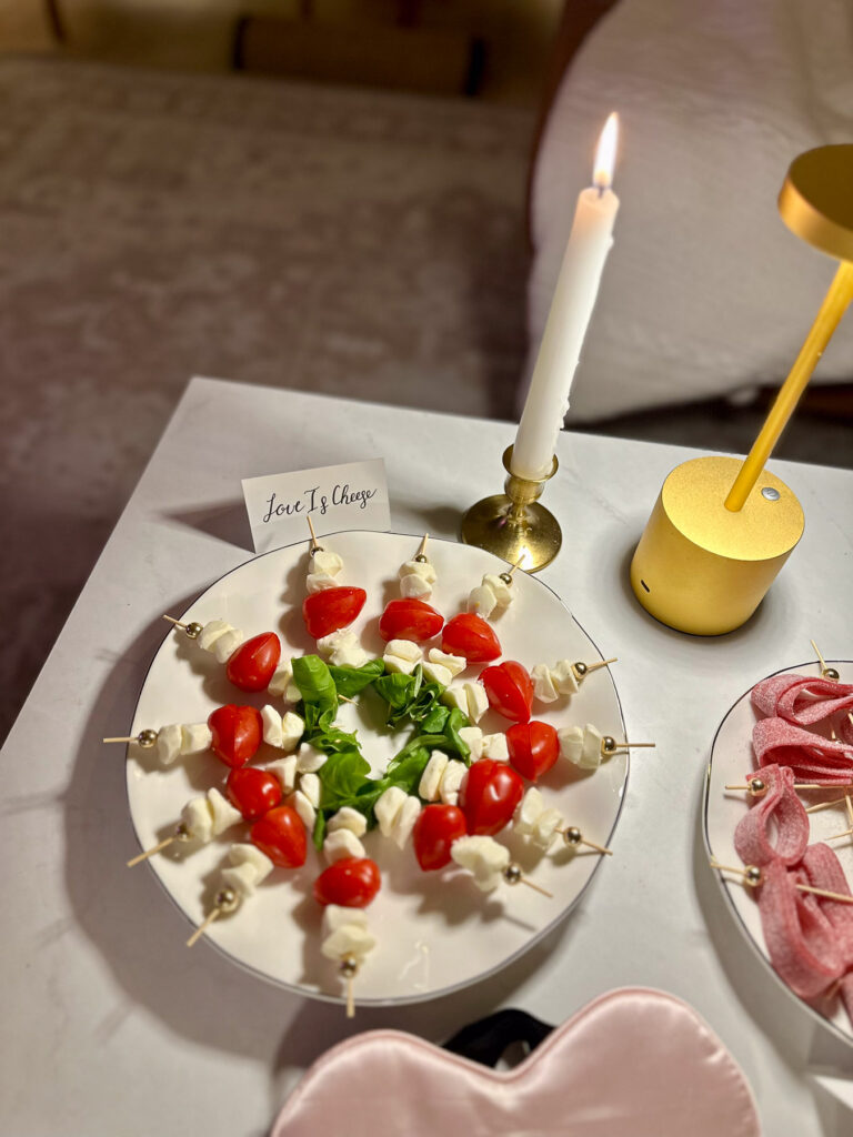 Caprese skewers arranged in a heart shape with a sign reading “Love Is Cheese” and gold decor on a marble table.
