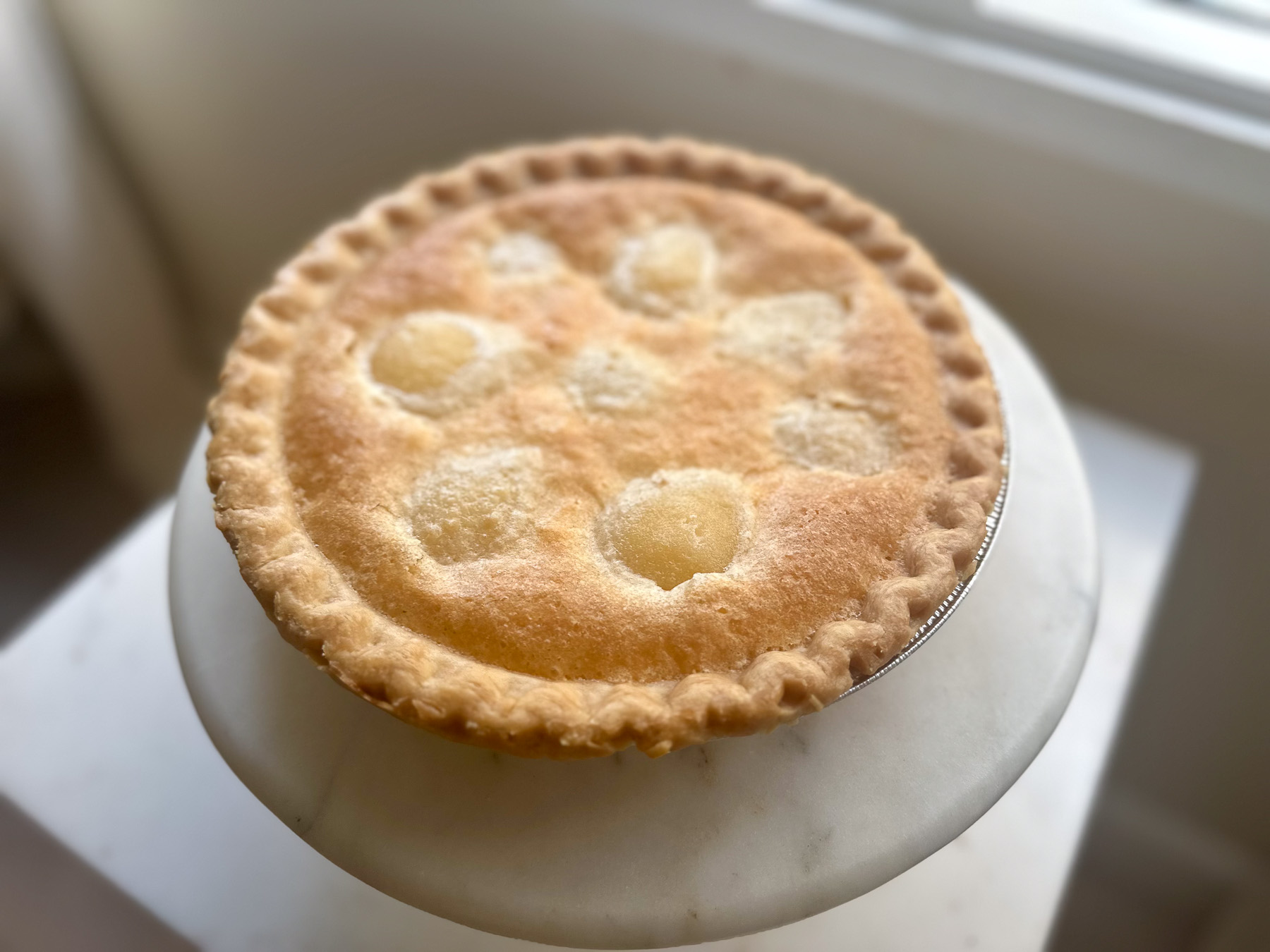 Golden brown old-fashioned pear pie with a buttery crust and baked pear pieces showing through the top, displayed on a marble cake stand near a window.