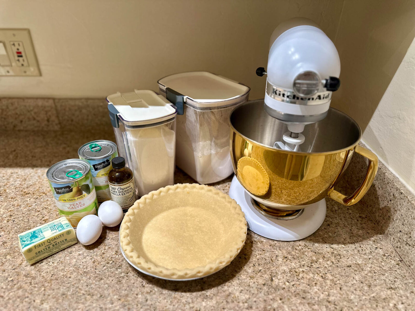 Ingredients for old-fashioned pear pie laid out beside a gold KitchenAid mixer — canned pears, butter, sugar, eggs, vanilla, and pie shell.