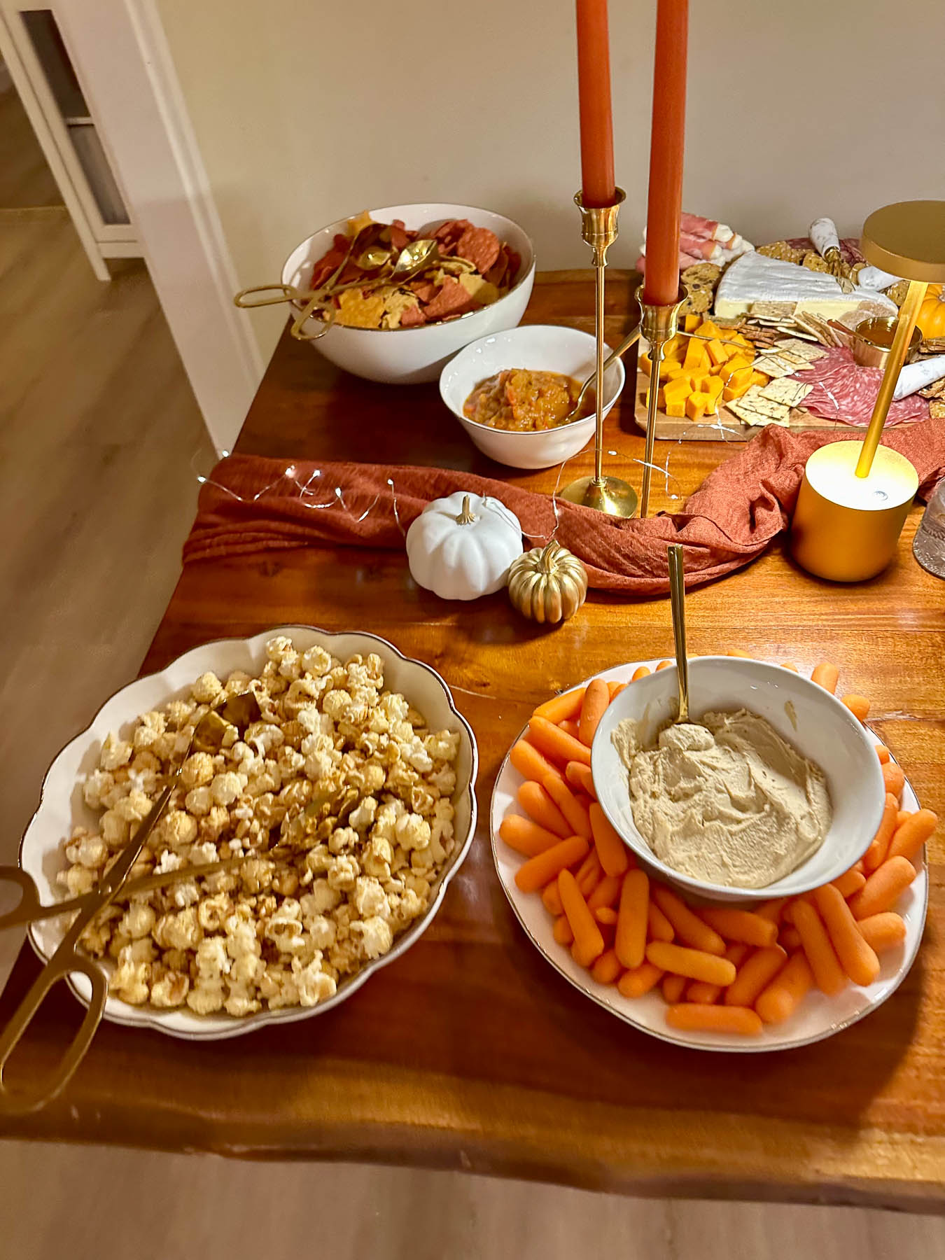 A festive fall snack spread on a wooden table with a white bowl of kettle corn, carrots with hummus, and a charcuterie board next to lit orange taper candles.