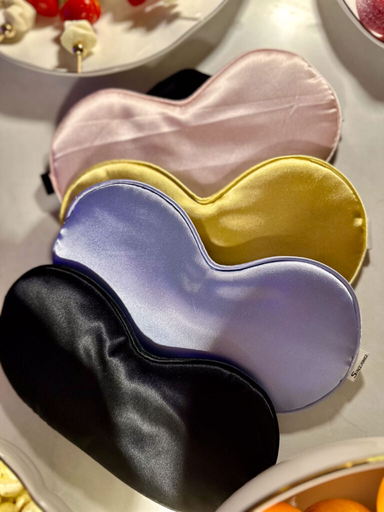 Four silk sleep masks in pastel pink, gold, lavender, and black displayed on a marble table for a Love Is Blind watch party.
