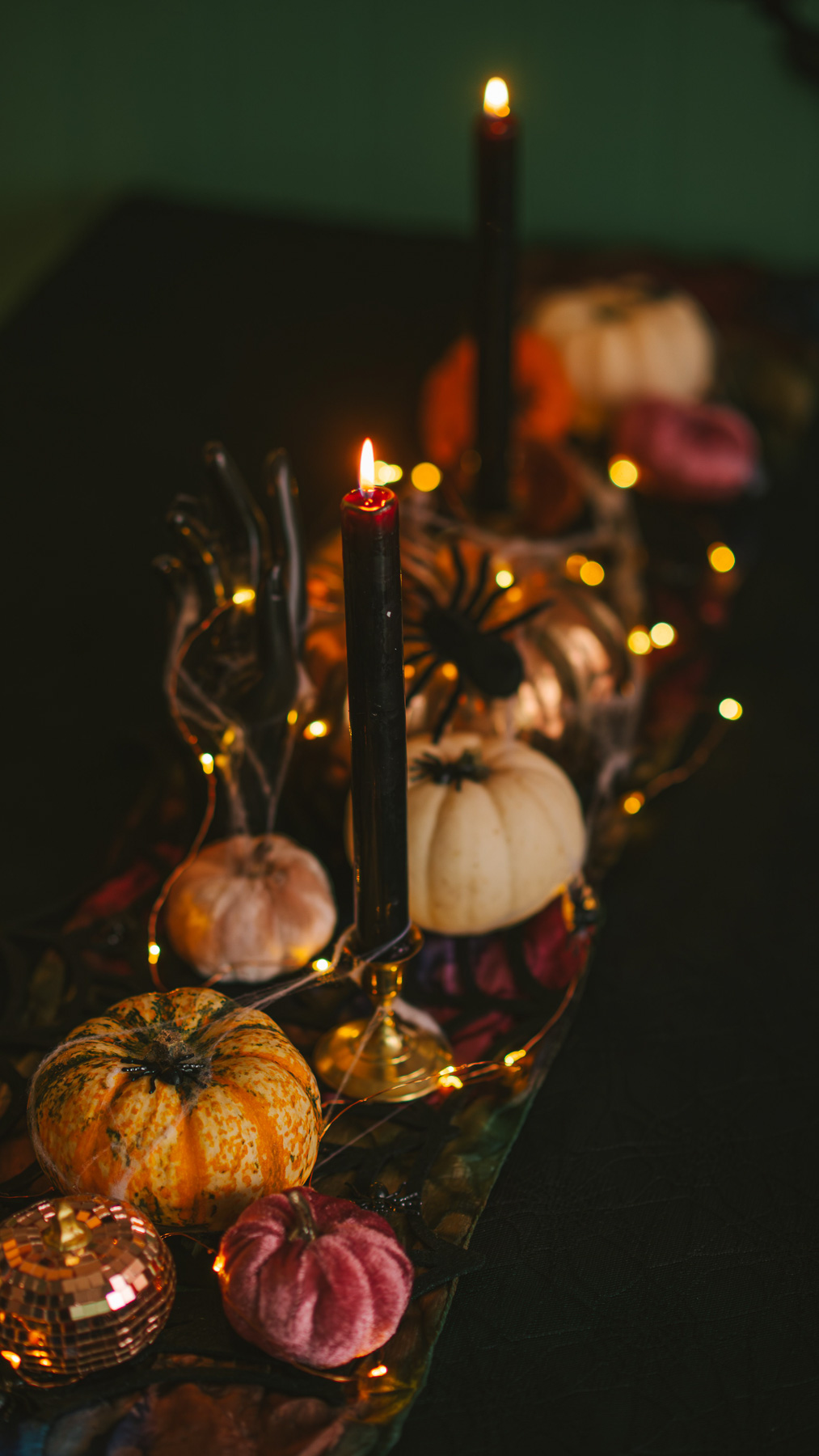 An elegant Spooky Glam Halloween tablescape featuring tall black taper candles, small white and orange pumpkins, a decorative spider, and warm fairy lights on a black tablecloth.