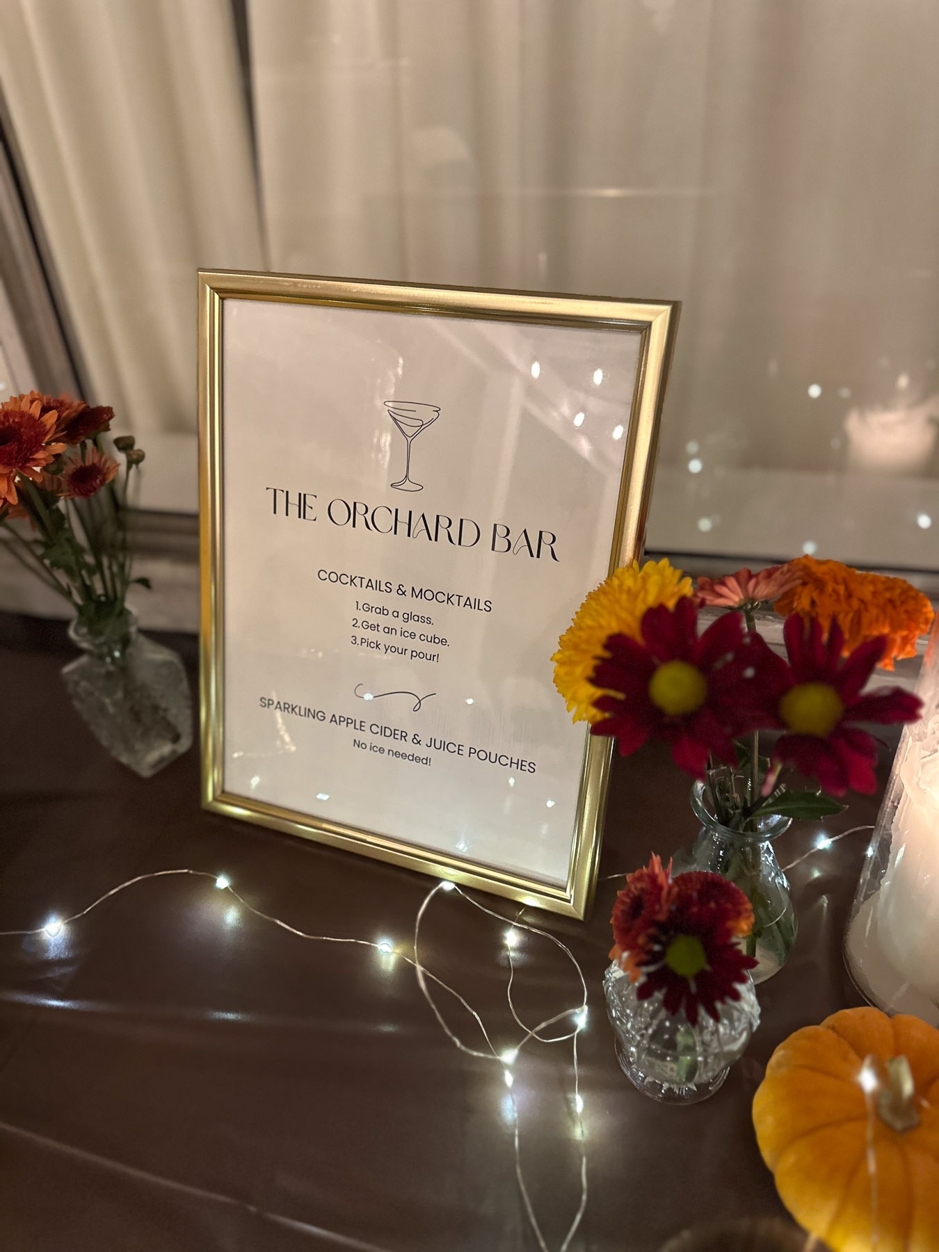 A close-up of a framed 'The Orchard Bar' drink menu on a table decorated with orange and burgundy flowers in glass vases, fairy lights, and a mini pumpkin.