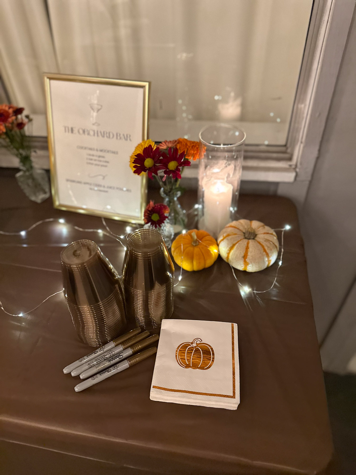 A cozy drink station on a brown tablecloth featuring stacked gold plastic cups, sharpie markers for labeling, a framed bar sign, and little decorative pumpkins wrapped in fairy lights.