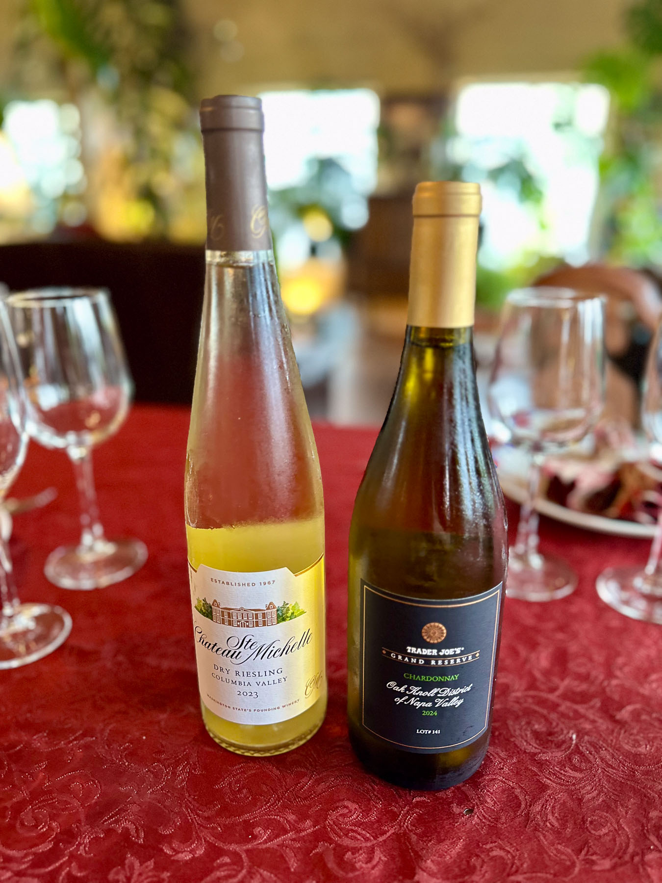 Close-up of Trader Joe’s Grand Reserve chardonnay and Chateau Ste. Michelle dry riesling bottles side by side on a red tablecloth.
