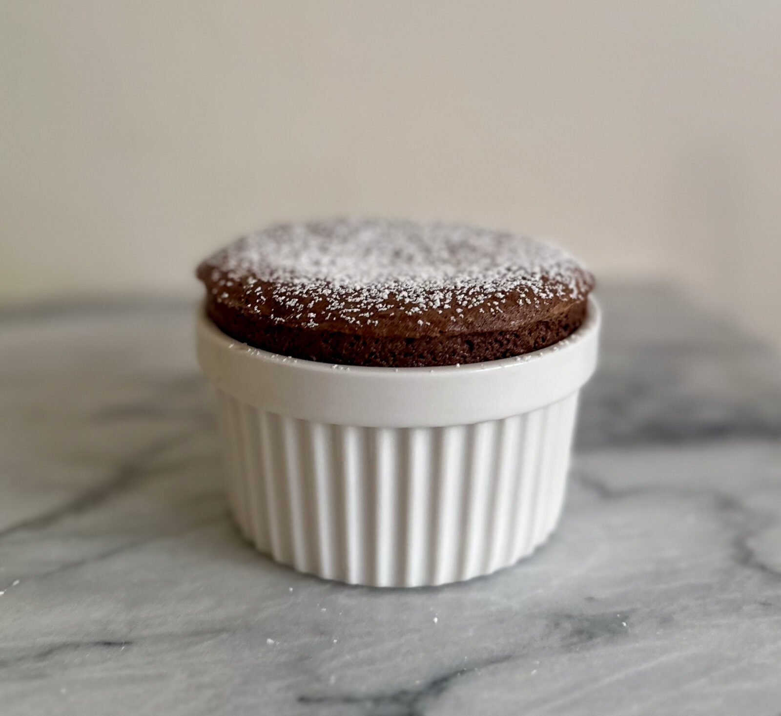 Individual chocolate soufflé dusted with powdered sugar in a white ramekin on a marble surface.