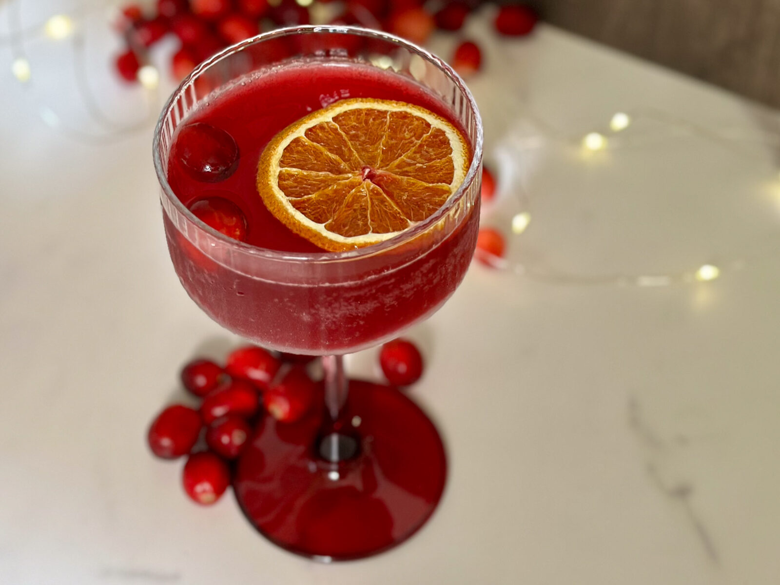 A deep red cranberry-orange mocktail inspired by the Cosmopolitan, served in a coupe glass with a dried orange slice and fresh cranberries.