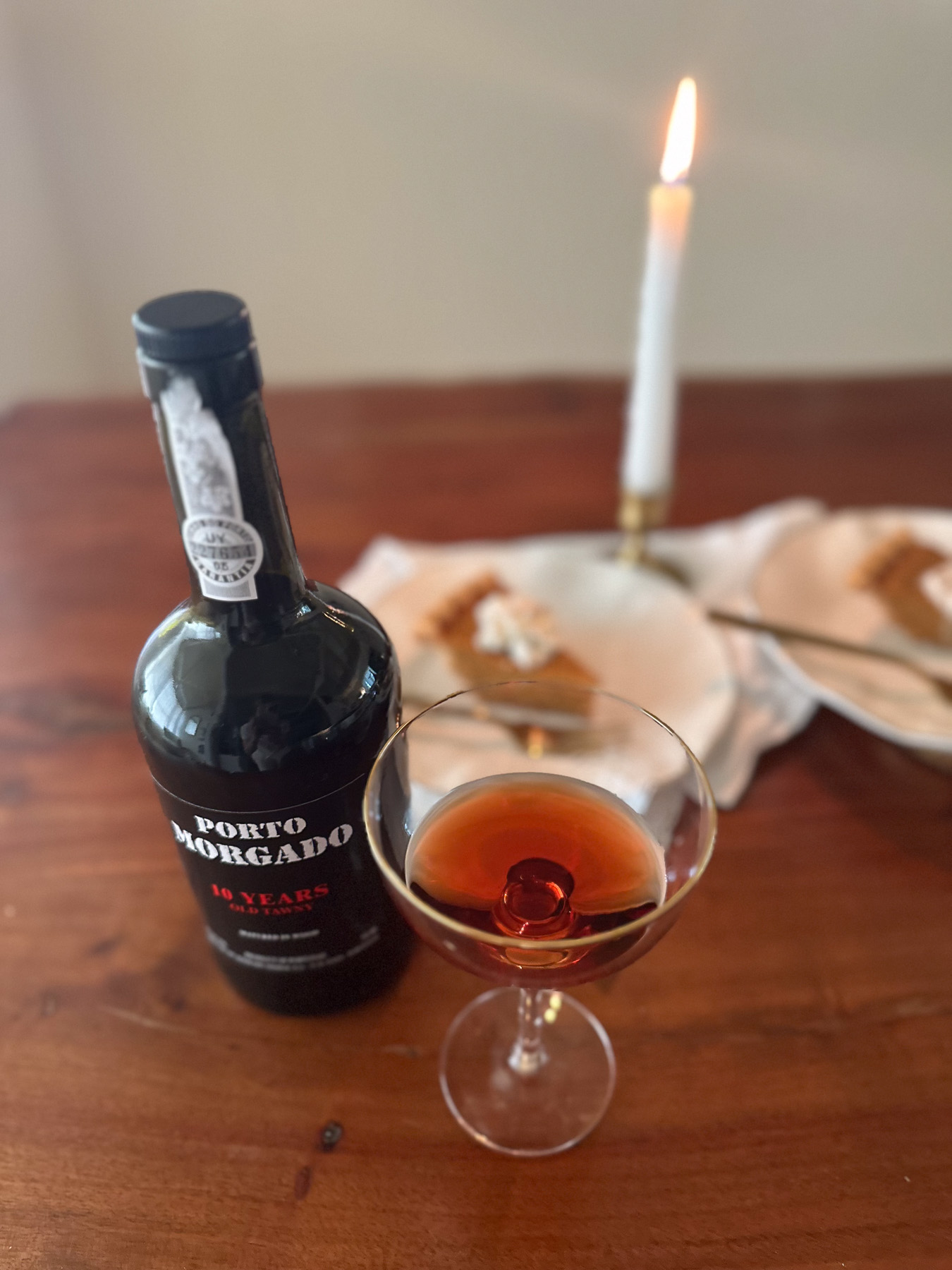 Glass of tawny port next to Porto Morgado bottle and pumpkin pie slices on a wooden table with a lit candle.