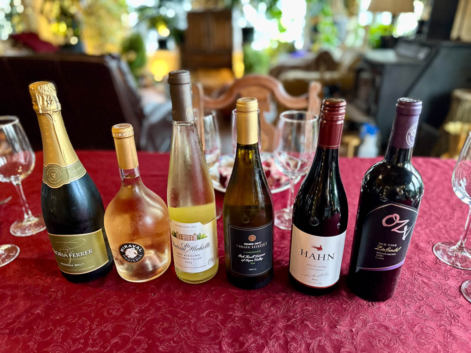 Overhead view of Thanksgiving wine tasting setup with a variety of bottles on a red tablecloth, including sparkling, white, rosé, and red wines arranged in a row.