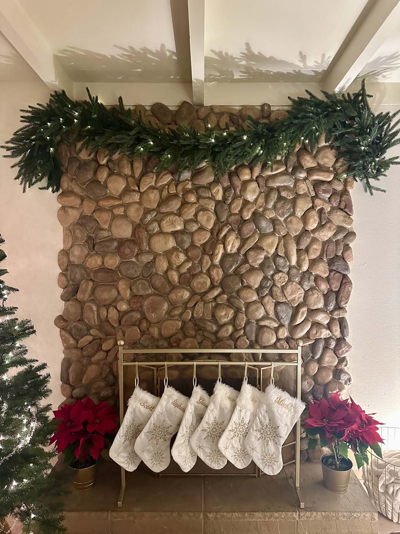 White and gold Christmas stockings hanging from a freestanding gold stocking stand in front of a stone fireplace with no mantel