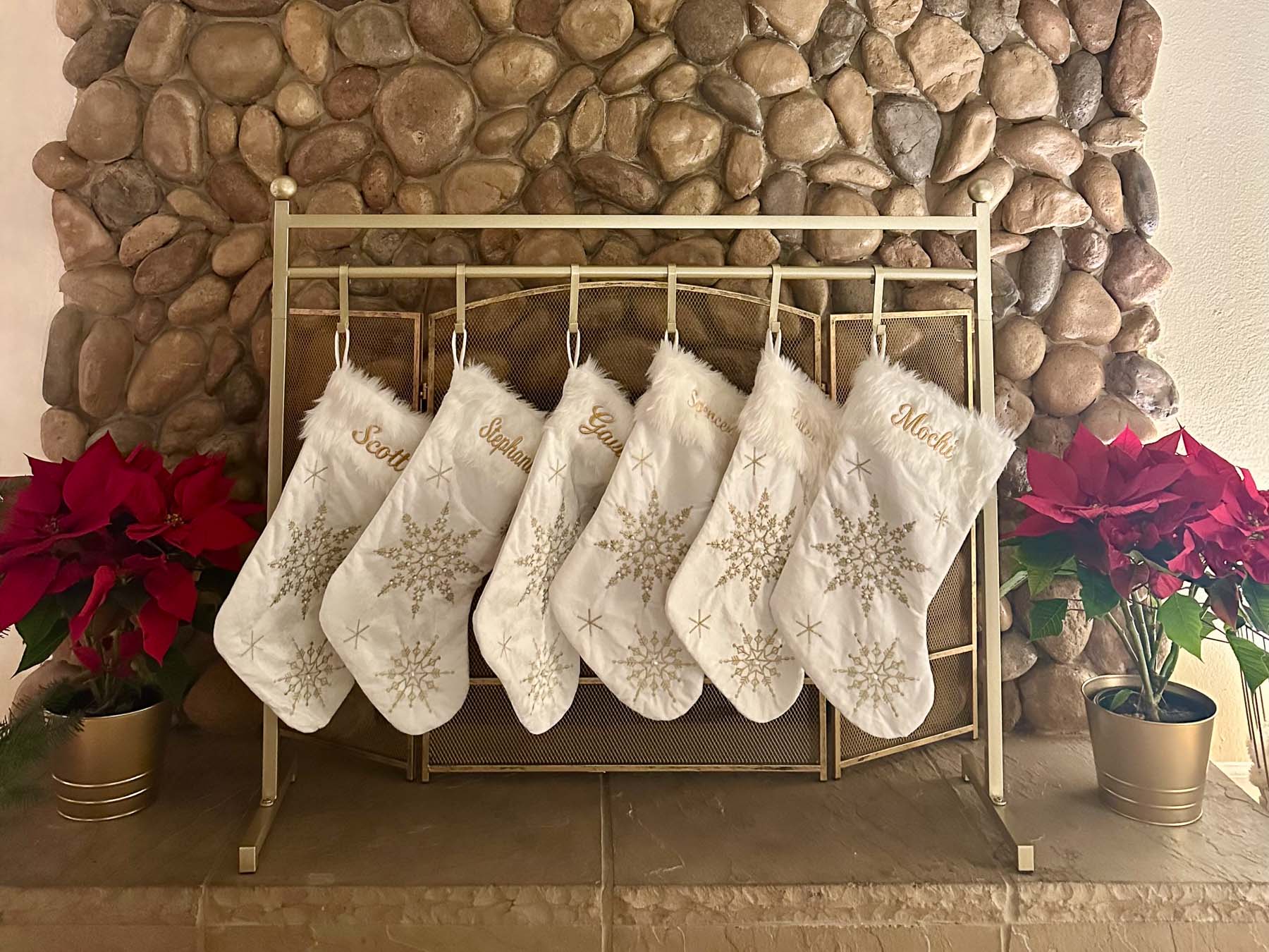 White and gold Christmas stockings hanging from a freestanding gold stocking stand in front of a stone fireplace without a mantel