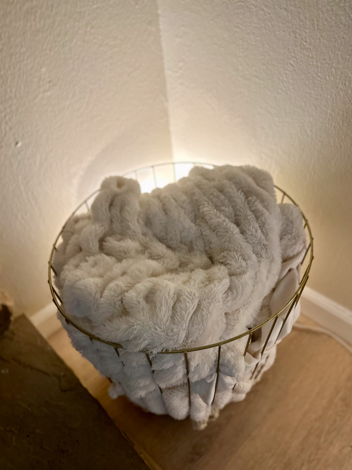 A plush ivory throw blanket nestled in a gold wire basket, softly lit by warm light in a corner nook.
