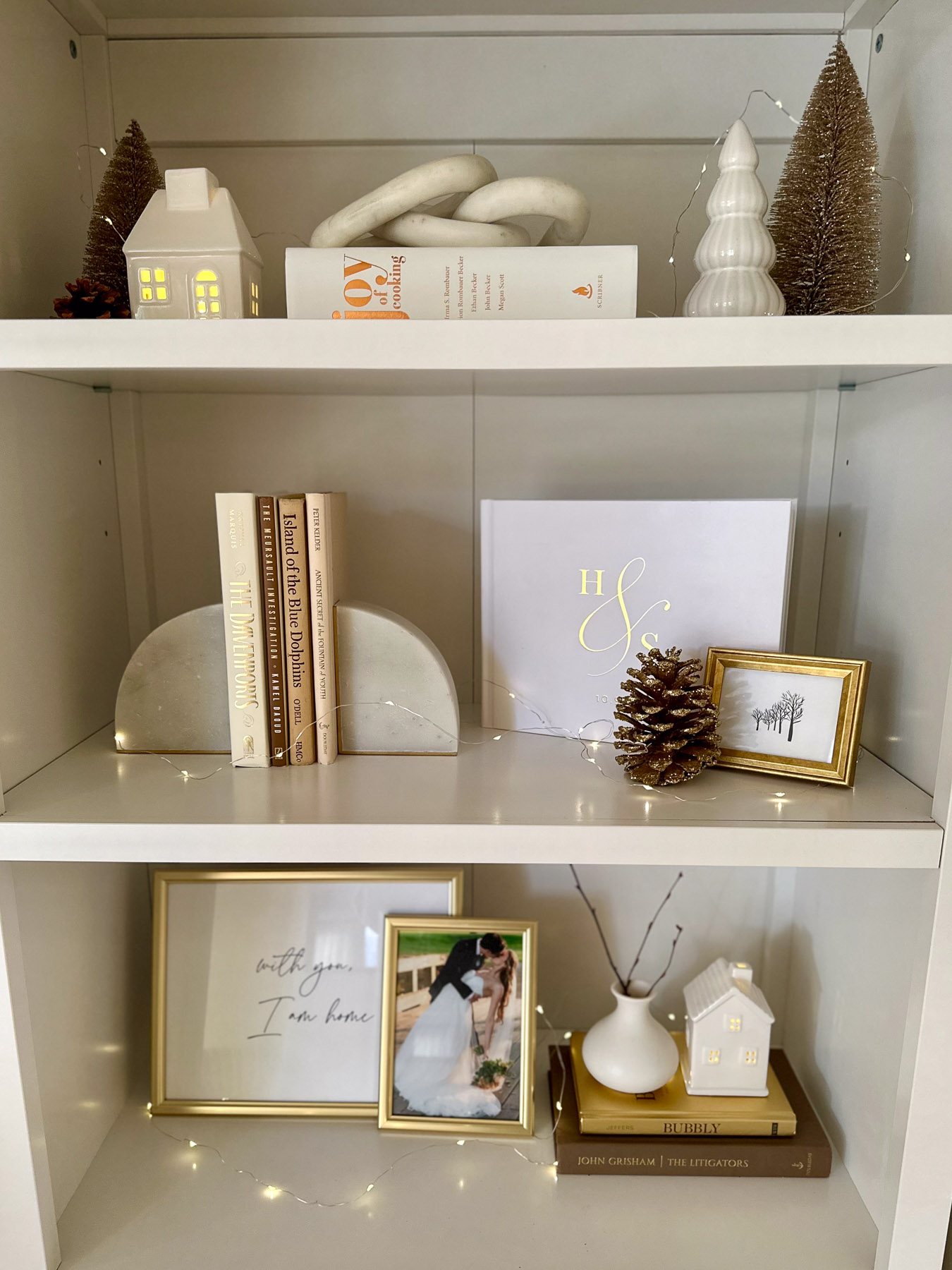 A white bookshelf decorated with neutral holiday items including ceramic houses, bottle brush trees, books, pinecones, and framed wedding photos. String lights are woven throughout for a cozy glow.