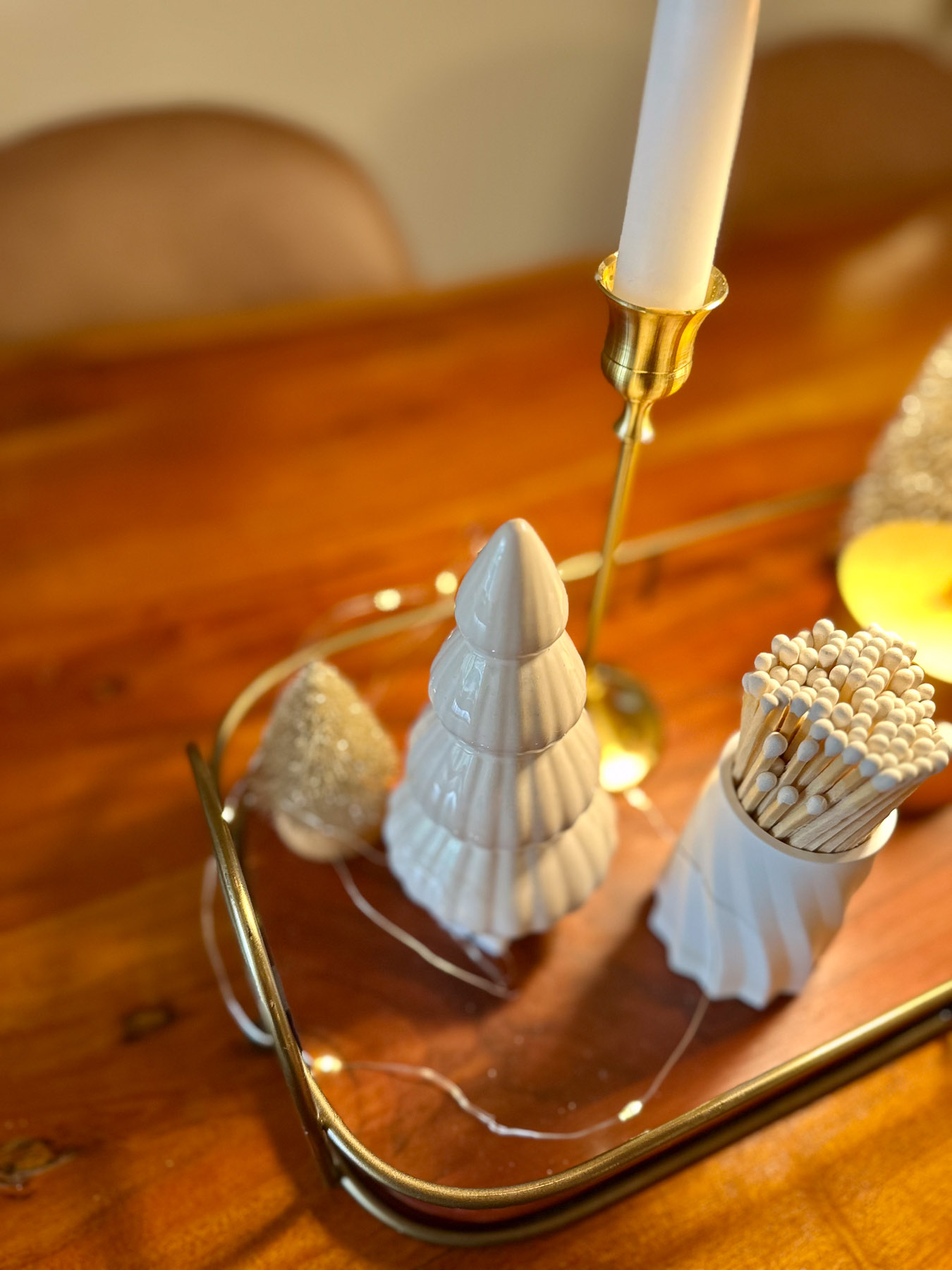 Close-up of a ceramic tree, glittered bottlebrush tree, and white matchstick holder arranged in a brass tray with twinkly lights.