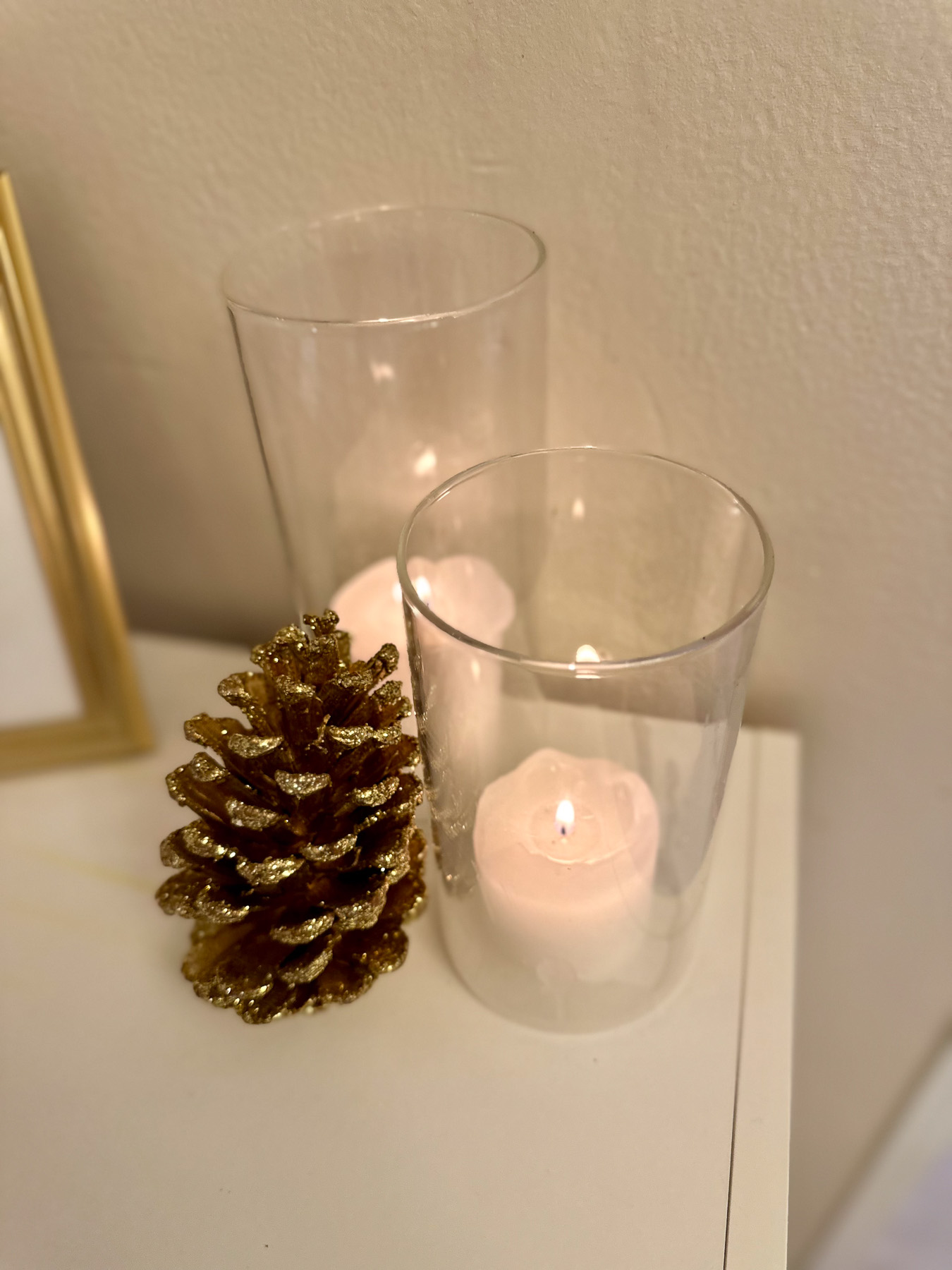 Gold-glittered pine cone and glass hurricane candles styled together on a white surface for a simple winter accent.