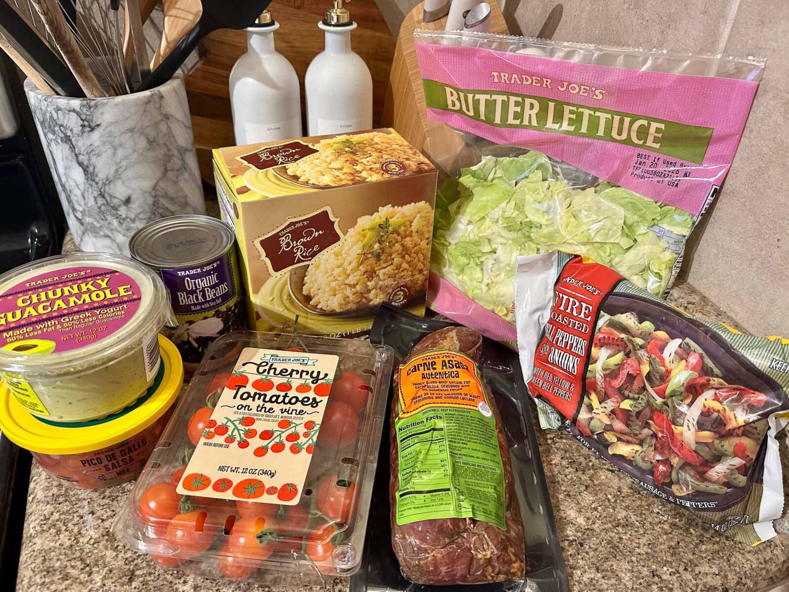 Trader Joe’s ingredients for carne asada bowls, including marinated carne asada, brown rice, butter lettuce, cherry tomatoes, pico de gallo, chunky guacamole, organic black beans, and fire-roasted peppers and onions.