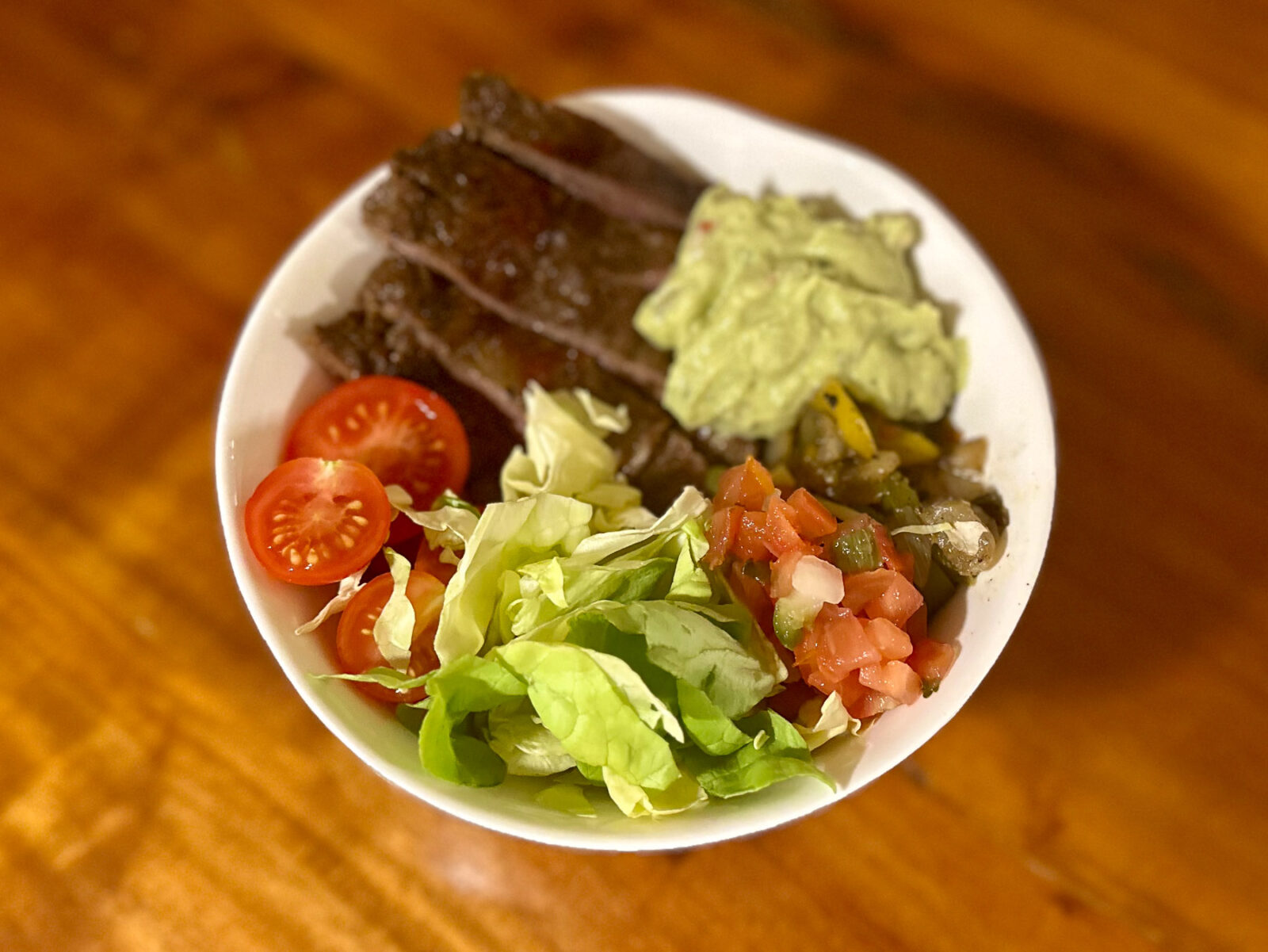 Carne asada bowl with sliced steak, cherry tomatoes, shredded lettuce, guacamole, fajita-style peppers and onions, and pico de gallo over brown rice.