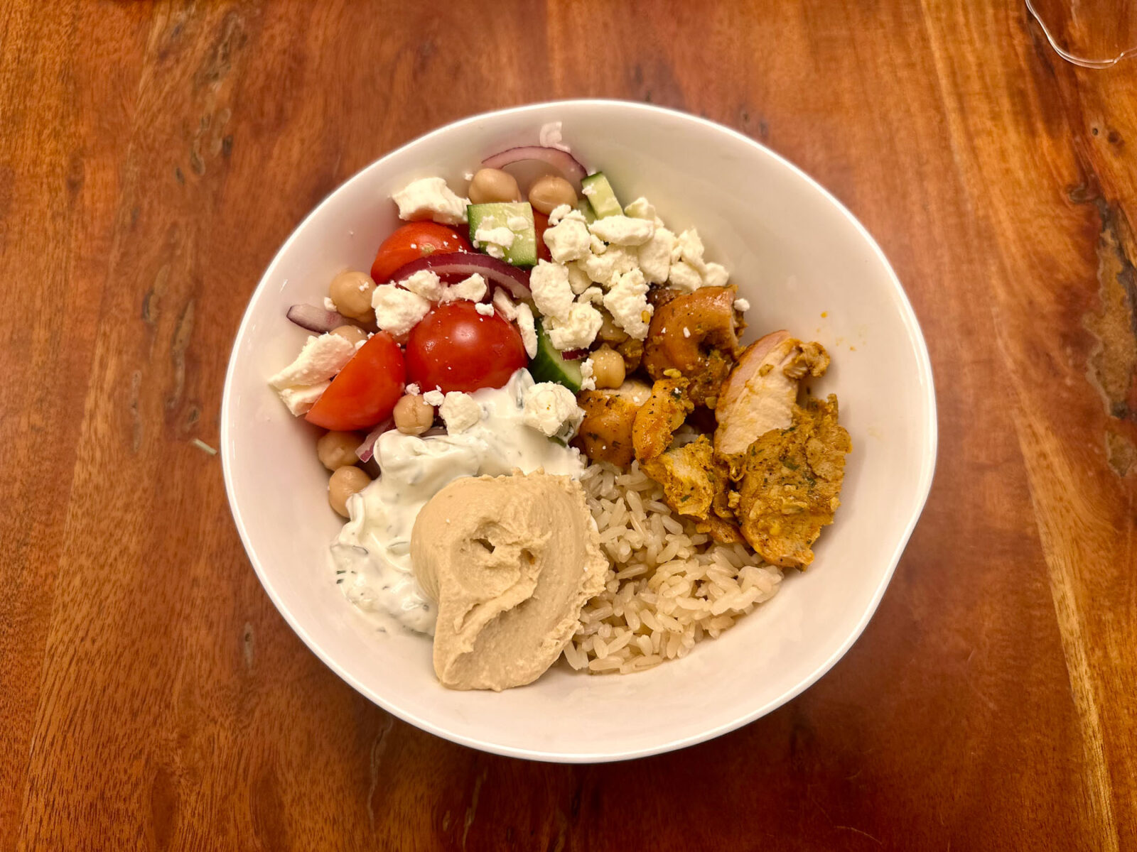Overhead view of a colorful Trader Joe’s chicken shawarma bowl with brown rice, marinated chicken, cherry tomato and cucumber salad, tzatziki, hummus, chickpeas, and feta.