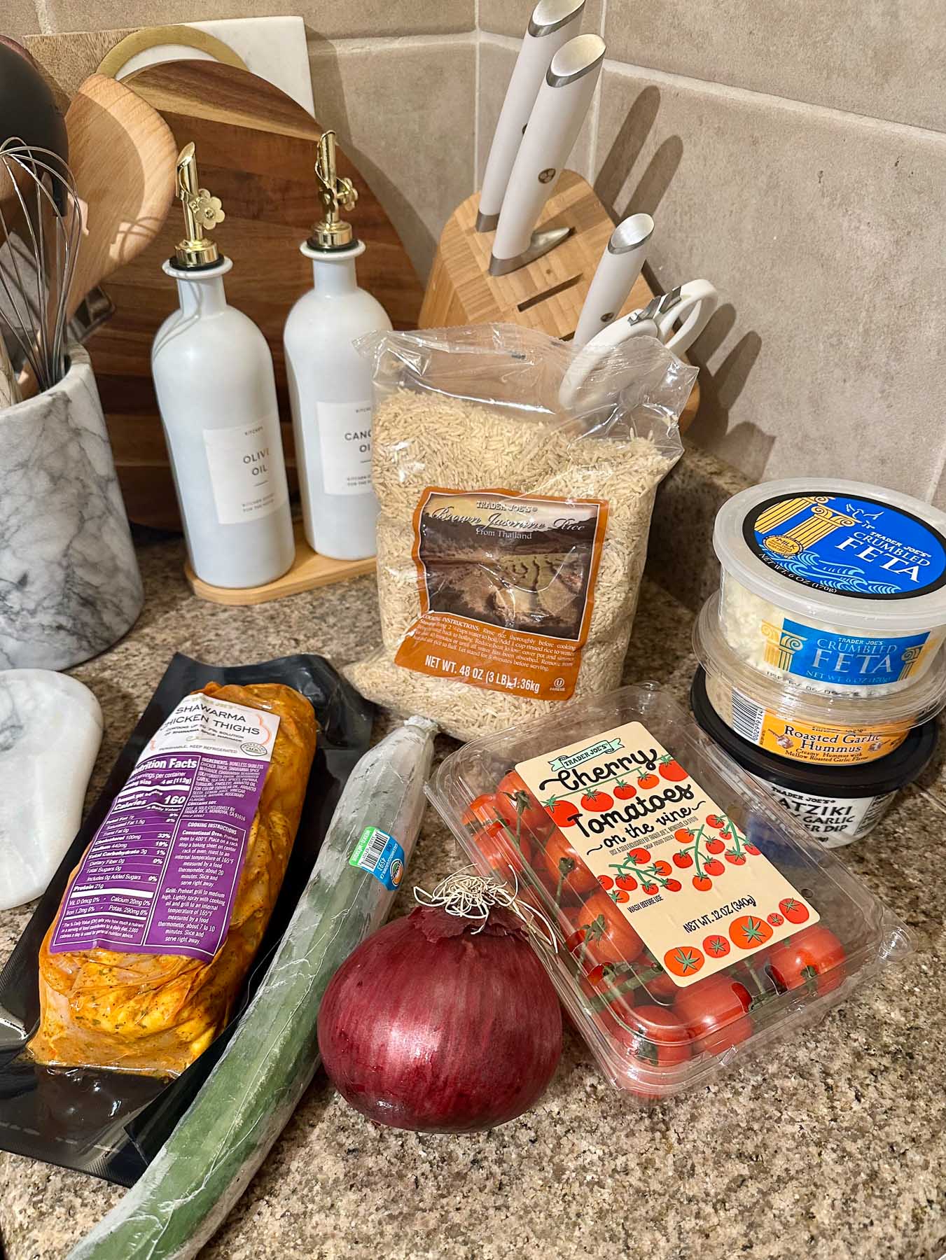 Trader Joe’s ingredients on a kitchen counter, including chicken shawarma, brown jasmine rice, cherry tomatoes, red onion, cucumber, crumbled feta, hummus, and tzatziki dip.