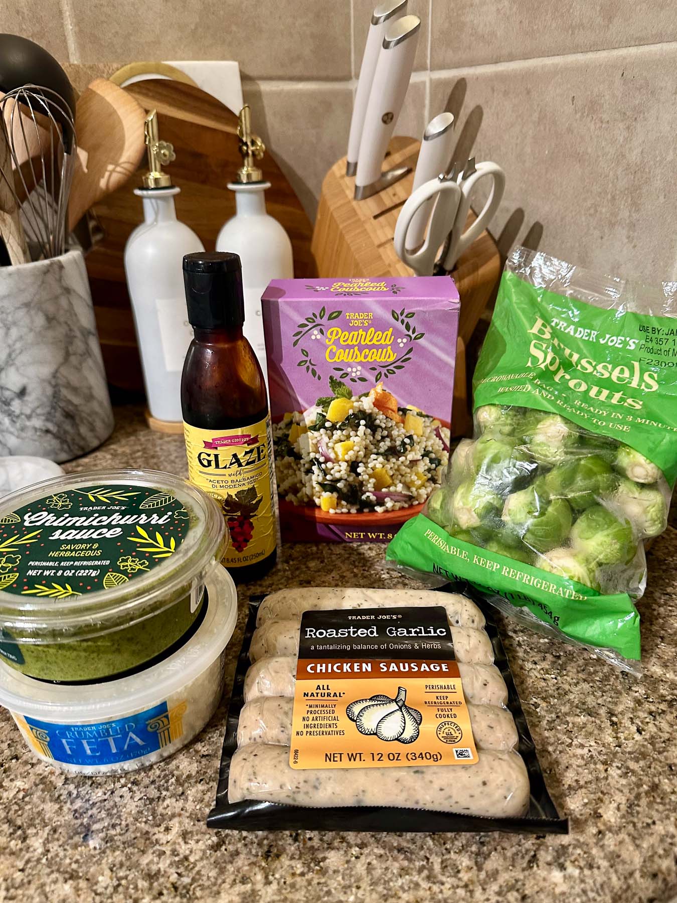 Trader Joe’s ingredients for a chimichurri chicken sausage bowl, including roasted garlic chicken sausage, Brussels sprouts, pearl couscous, chimichurri sauce, crumbled feta, and balsamic glaze.
