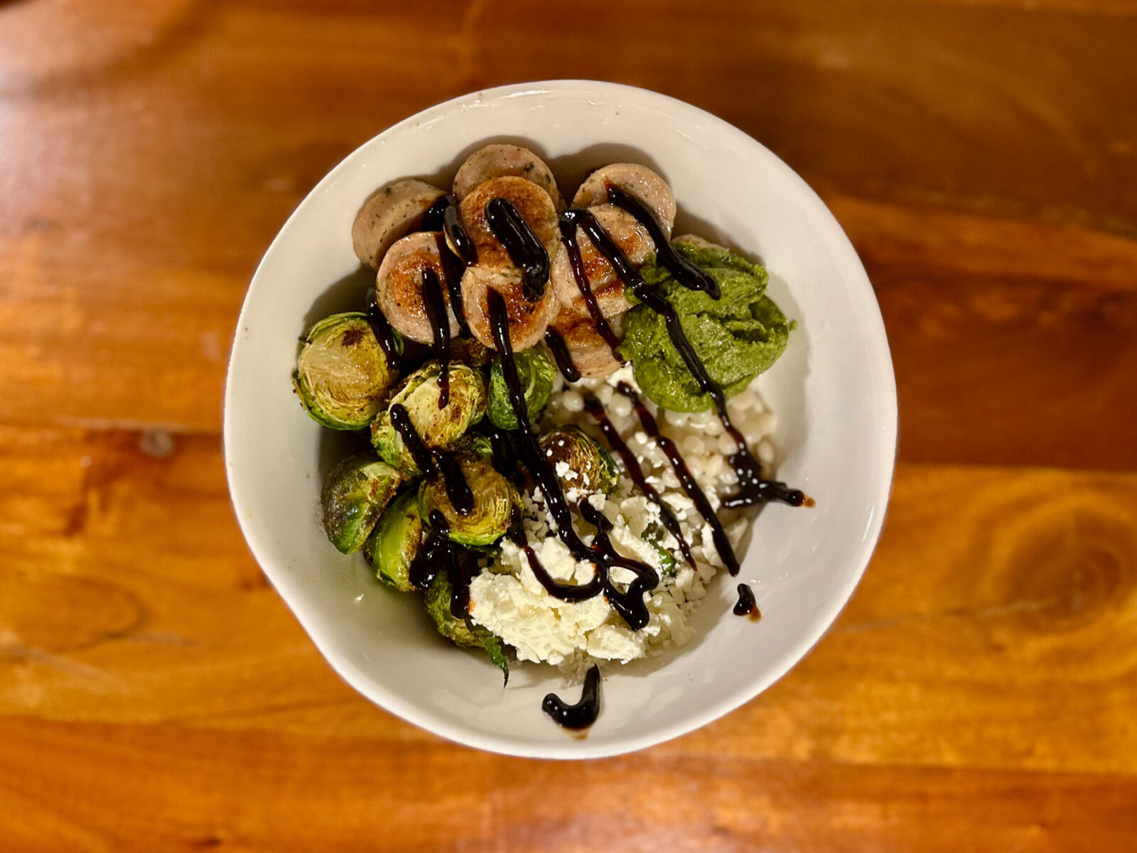 Overhead view of a bowl with roasted garlic chicken sausage, Brussels sprouts, chimichurri, pearl couscous, crumbled feta, and a drizzle of balsamic glaze.