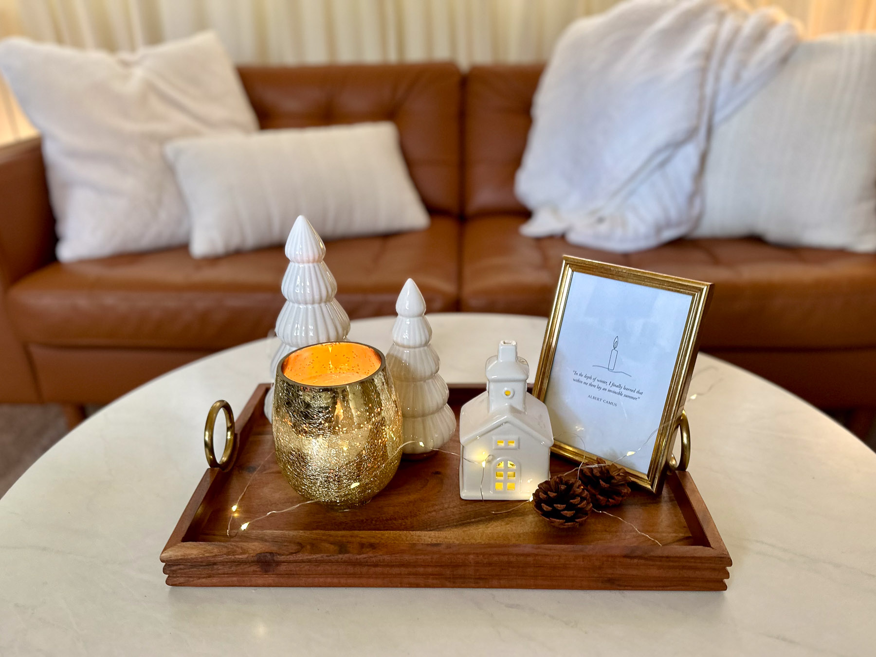 Winter console vignette styled on a wood tray with ceramic trees, a candle in a gold vessel, battery fairy lights, pine cones, and a framed minimalist winter quote.