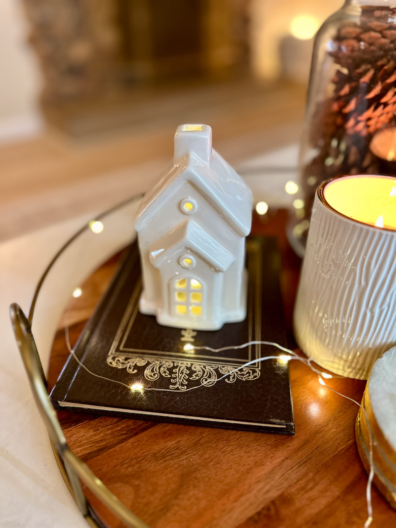 A ceramic house with lit windows sits on top of a brown and gold book, styled on a wooden tray with a candle, pinecones, and string lights.