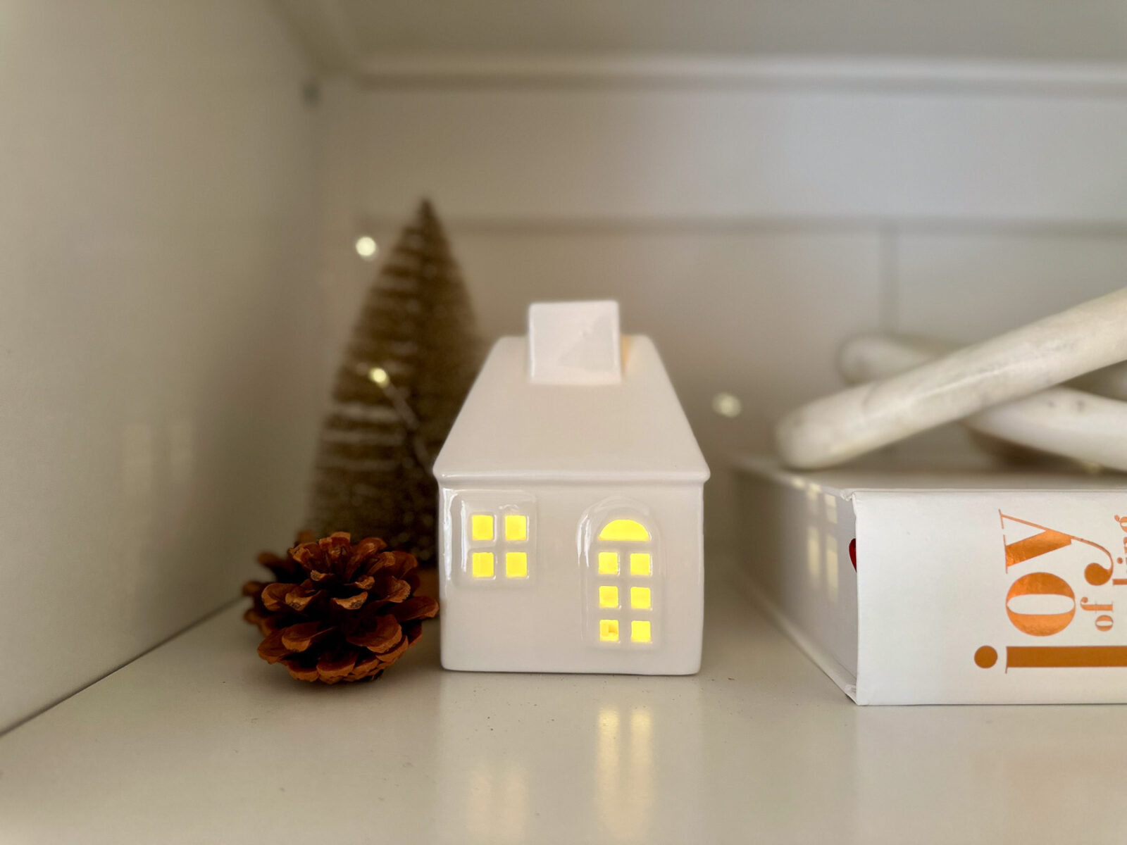 Close-up of a white ceramic house with glowing windows, nestled beside a pinecone and neutral-toned bottlebrush tree with twinkle lights.