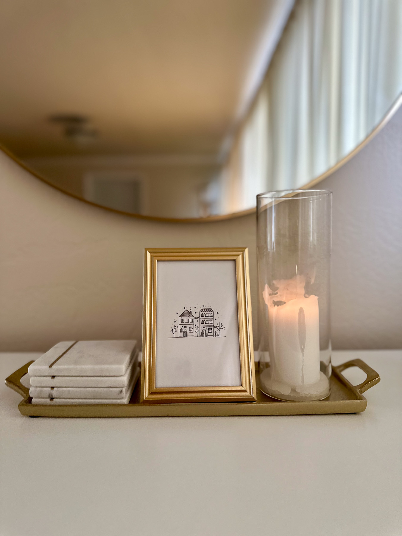 A rectangular gold tray on a white console holds one gold-framed winter-themed line drawing, stacked coasters, and a tall hurricane candle with warm lighting reflected in a nearby mirror.
